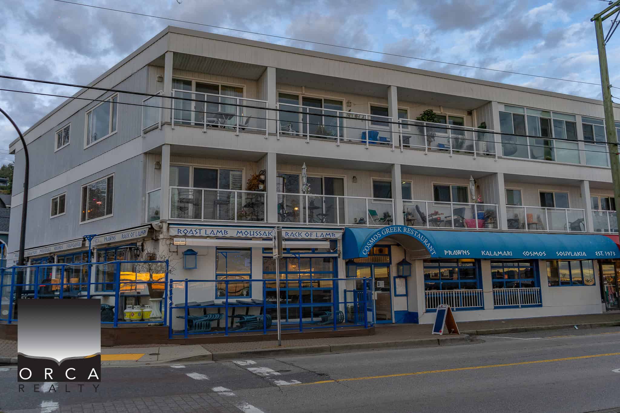 Modern multi-story building with oceanfront Greek restaurant on ground floor and residential units above, located on a busy street in Vancouver, BC, showcasing vibrant local dining and urban living options.
