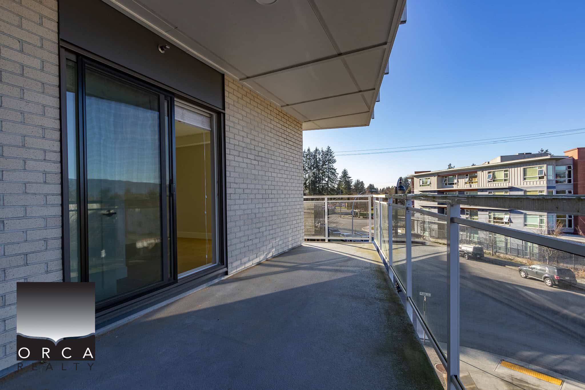 Balcony view from a modern apartment at Orca Realty Inc. showcasing a spacious outdoor balcony, glass railing, and cityscape setting with residential buildings nearby, perfect for urban living in Canada.
