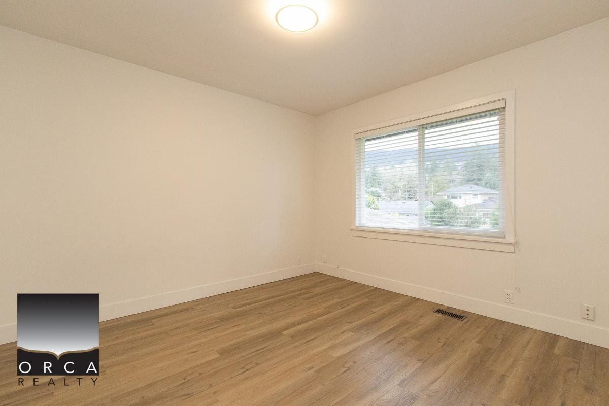 Modern empty apartment room with white walls, large window with blinds, and wooden flooring, showcasing Orca Realty's property listings and real estate services in British Columbia.