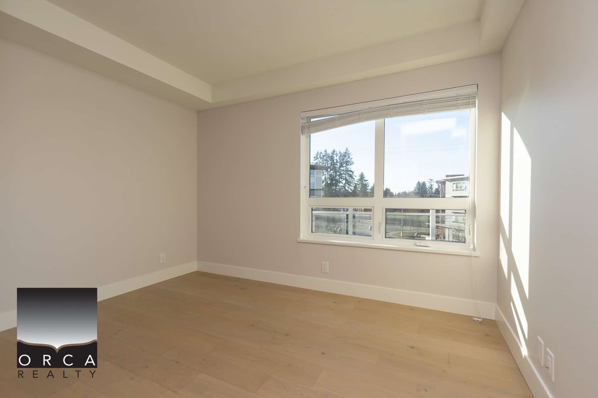 Bright, modern empty bedroom with large window, hardwood flooring, white walls, and natural sunlight, ideal for Vancouver real estate buyers seeking stylish condos or apartments.