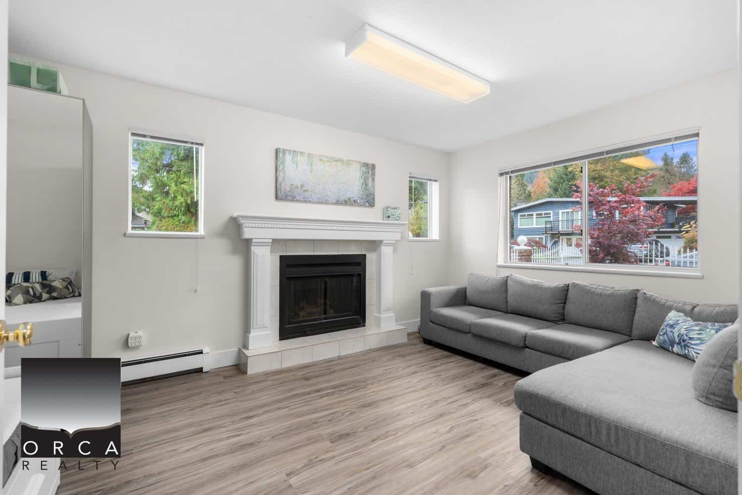 Modern living room with fireplace, large windows showcasing fall foliage, grey sectional sofa, hardwood floors, bright and welcoming space perfect for Vancouver Island homebuyers.