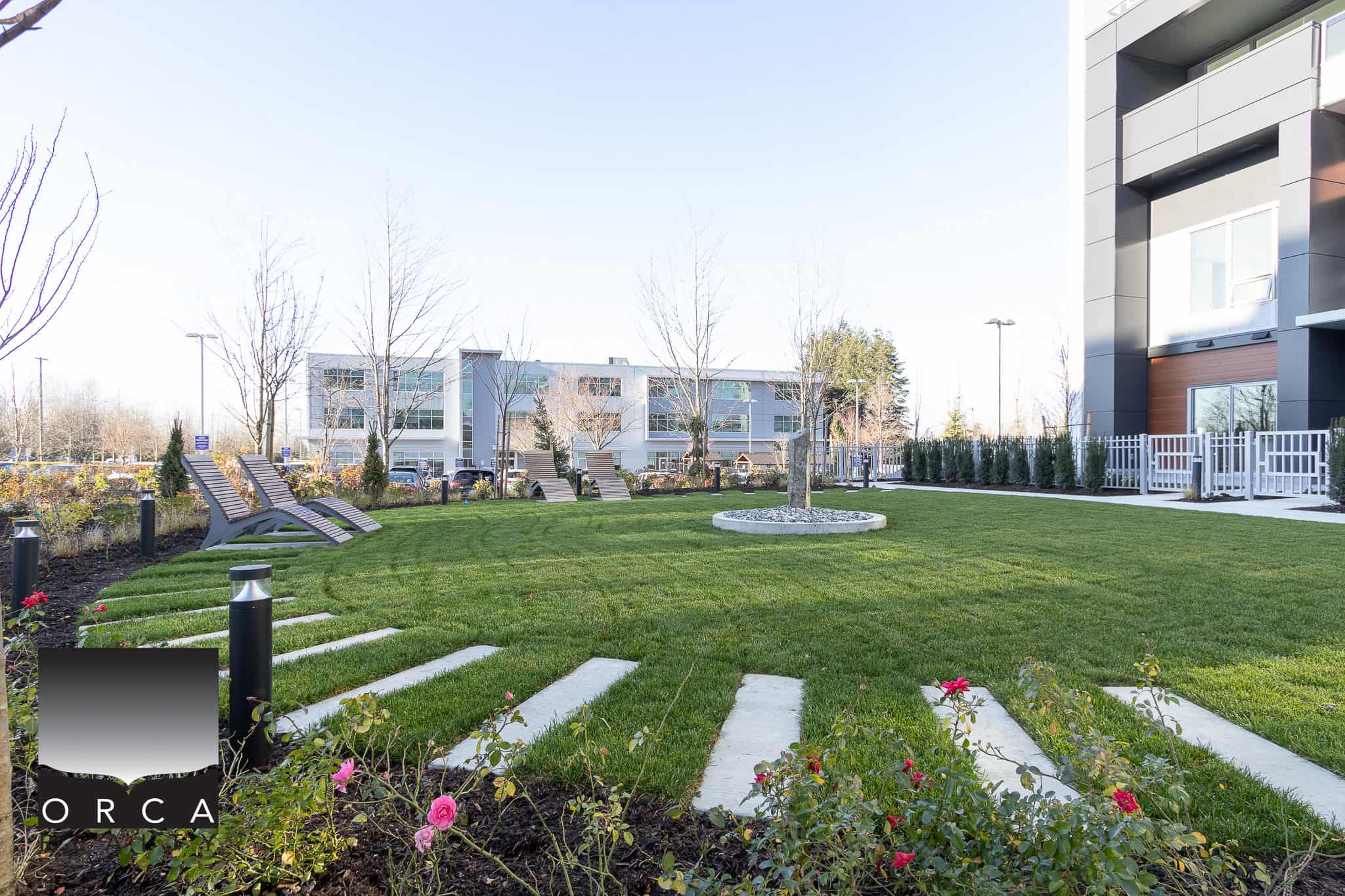 Lush green landscaped outdoor space with lounge chairs, young trees, and modern apartment buildings in the background, showcasing expert property management by Orca Realty Inc.