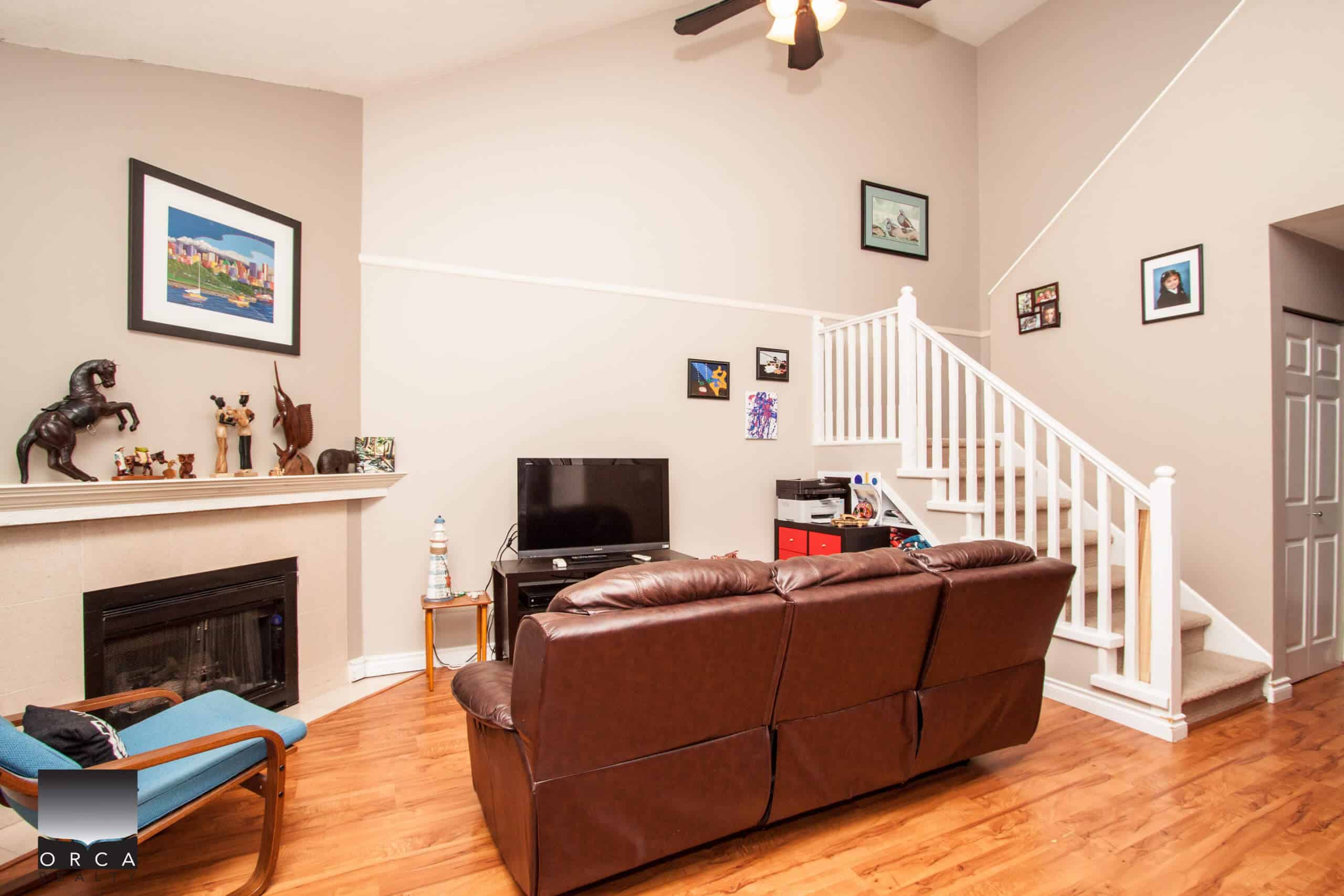 Cozy living room with a brown leather sofa, fireplace, wall art, and staircase, showcasing a comfortable home interior offered by Orca Realty Inc.