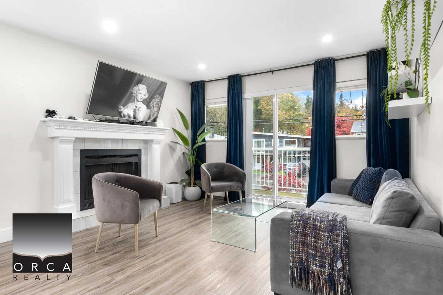Bright modern living room with large windows, cozy grey sofa, stylish chairs, a fireplace with a TV above, and elegant blue curtains, ideal for residential real estate in Vancouver.