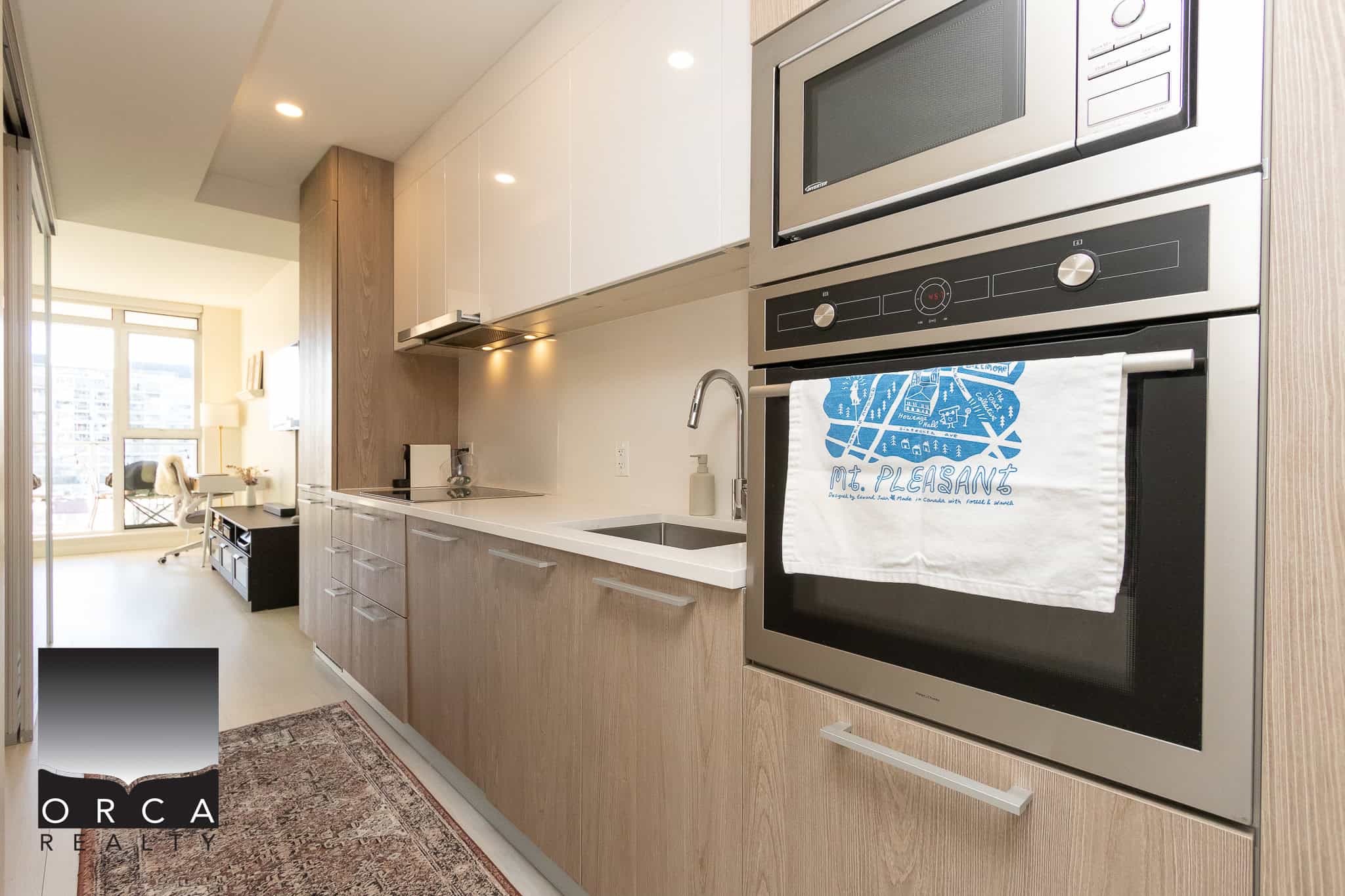 Modern Vancouver condominium kitchen with stainless steel appliances, white and wood cabinetry, and a view of a bright living space with large windows. Perfect for contemporary urban living.