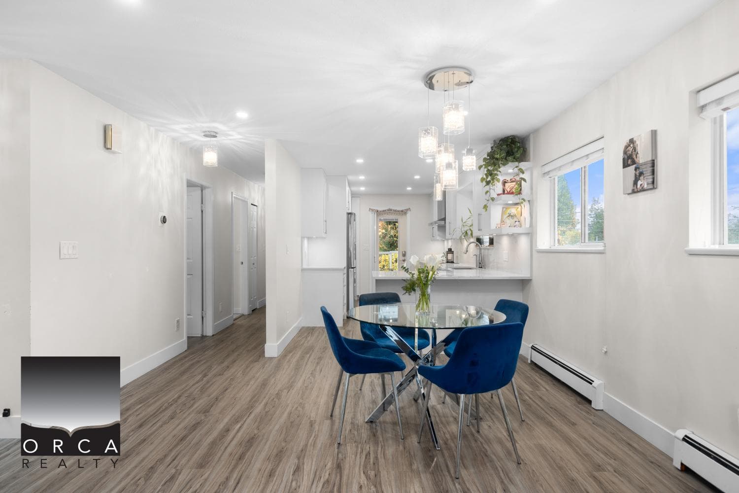Modern open-concept dining area in a contemporary home, featuring blue velvet chairs, a glass table, and a bright, airy kitchen with large windows and stylish pendant lighting, ideal for family living and entertaining.