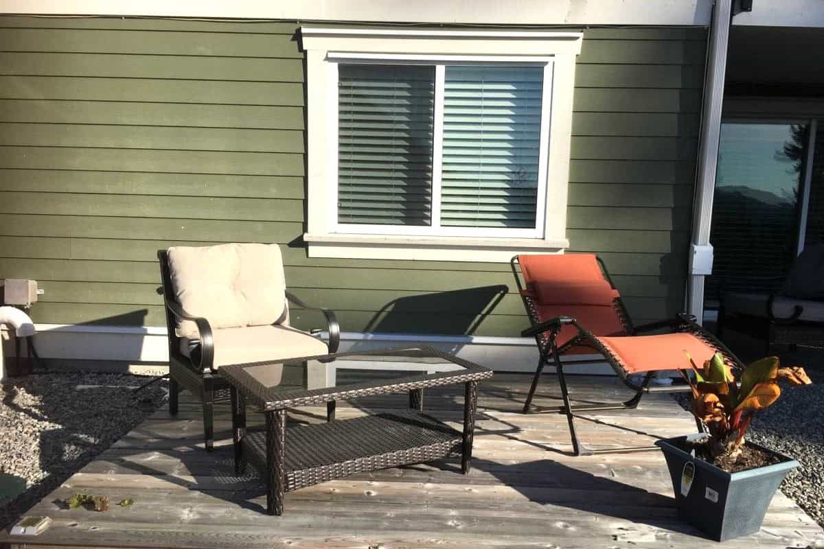 Outdoor patio with seating area featuring a beige cushioned chair, orange lounge chair, glass-top table, and potted plant in front of a green house exterior with a window.