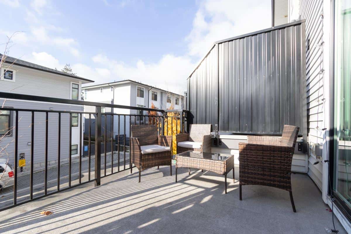 Spacious outdoor balcony with wicker chairs, glass table, and modern black railing overlooking residential townhomes in Victoria, BC. Perfect for relaxing or entertaining.
