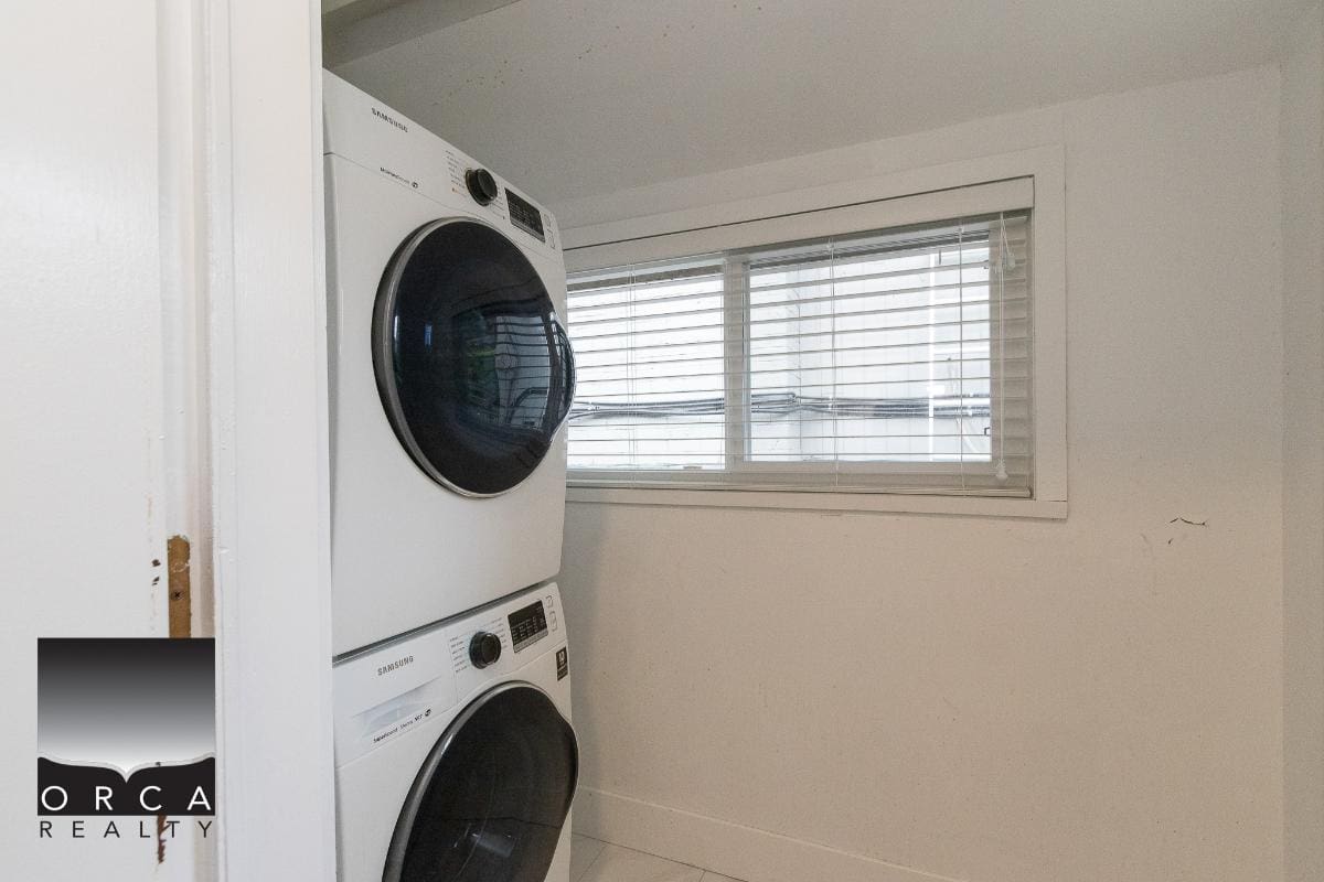 Front-loading washer and dryer stacked in a bright laundry room with window blinds at Orca Realty Inc. Vancouver Island real estate.