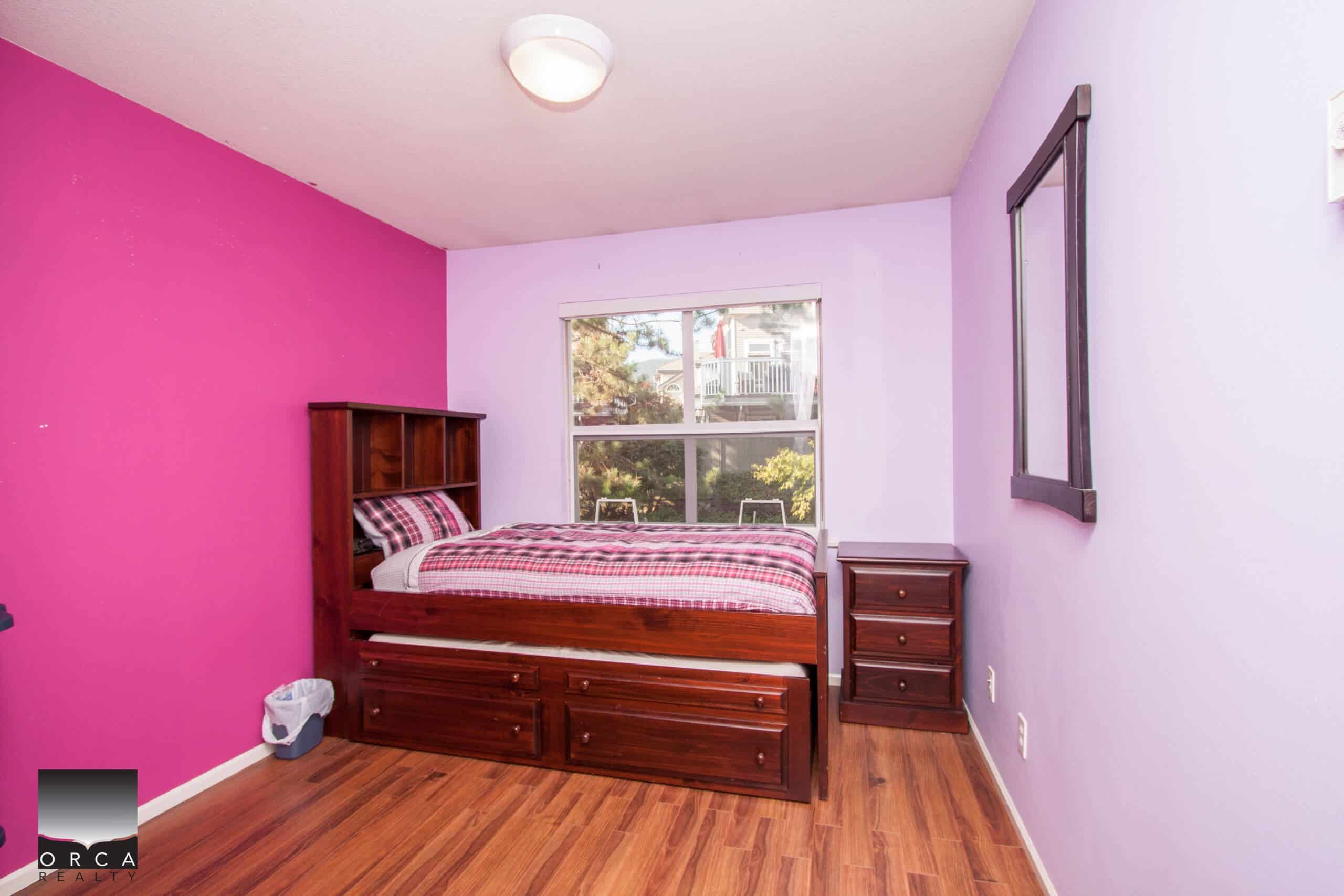 Bright bedroom with vibrant pink and purple painted walls, wooden flooring, large window providing natural light, and a cozy bed with matching wooden furniture, perfect for a comfortable home.