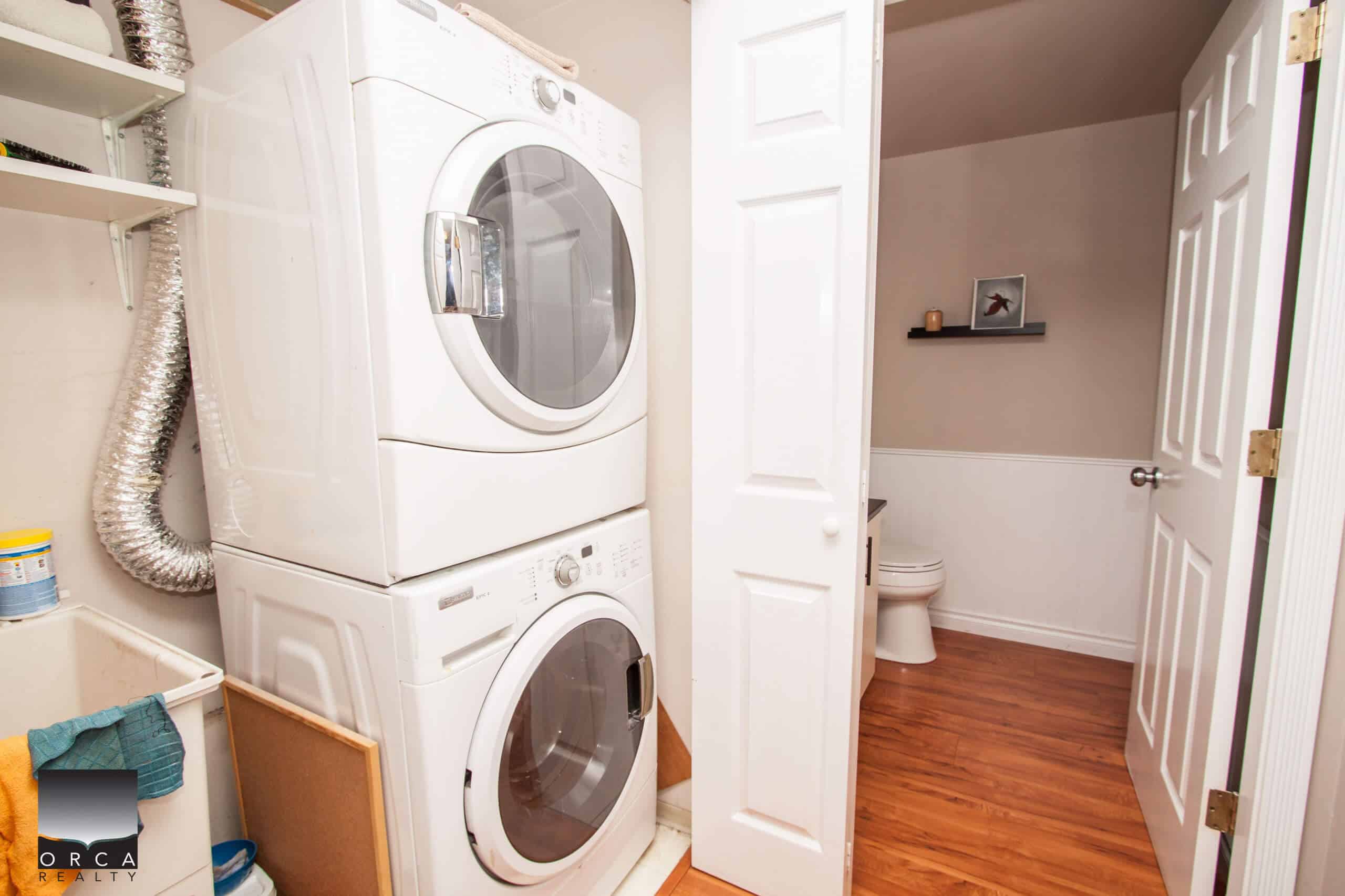 Stacked washer and dryer in laundry room with nearby bathroom, hardwood flooring, beige walls, and modern decor, showcasing efficient home laundry and bathroom features.