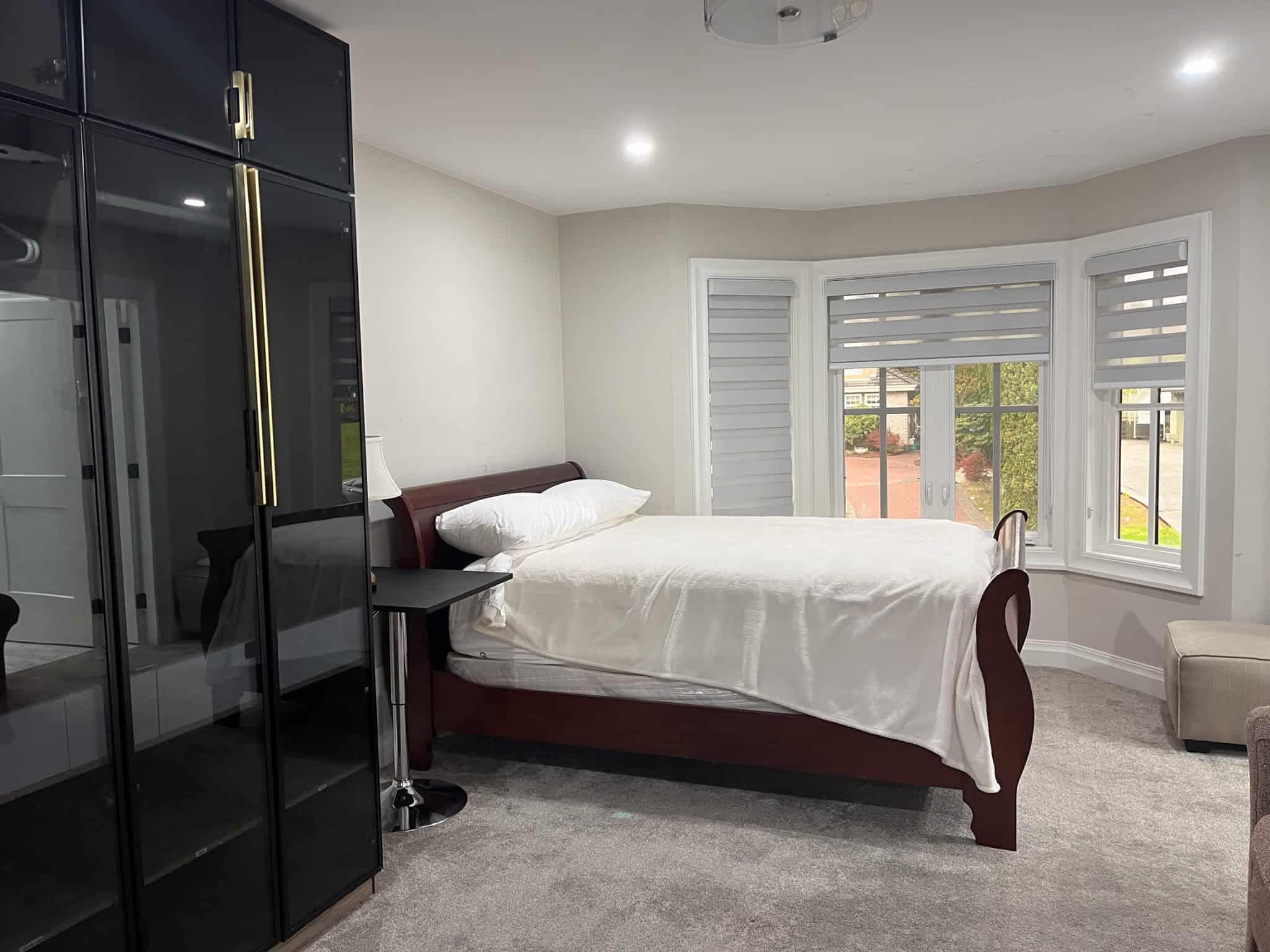 Modern bedroom with large bay windows, white walls, and plush carpet, featuring a dark wood bed frame, black wardrobe, and cozy seating area, perfect for Vancouver Island living.