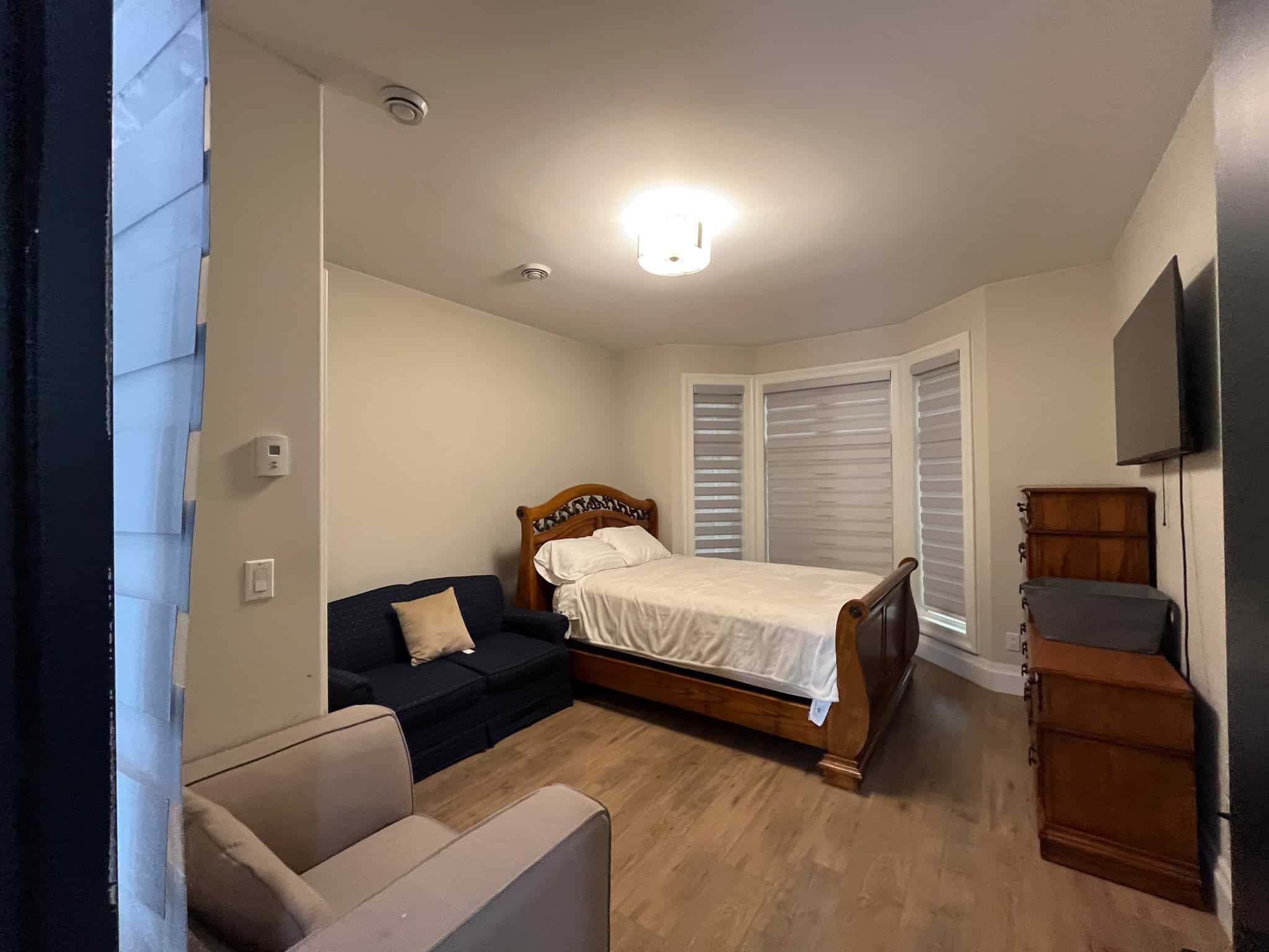 Modern bedroom with wood flooring, large bay windows with blinds, wooden bed frame, navy sofa, beige armchair, mounted TV, and vintage chest of drawers, offering a cozy, stylish living space.