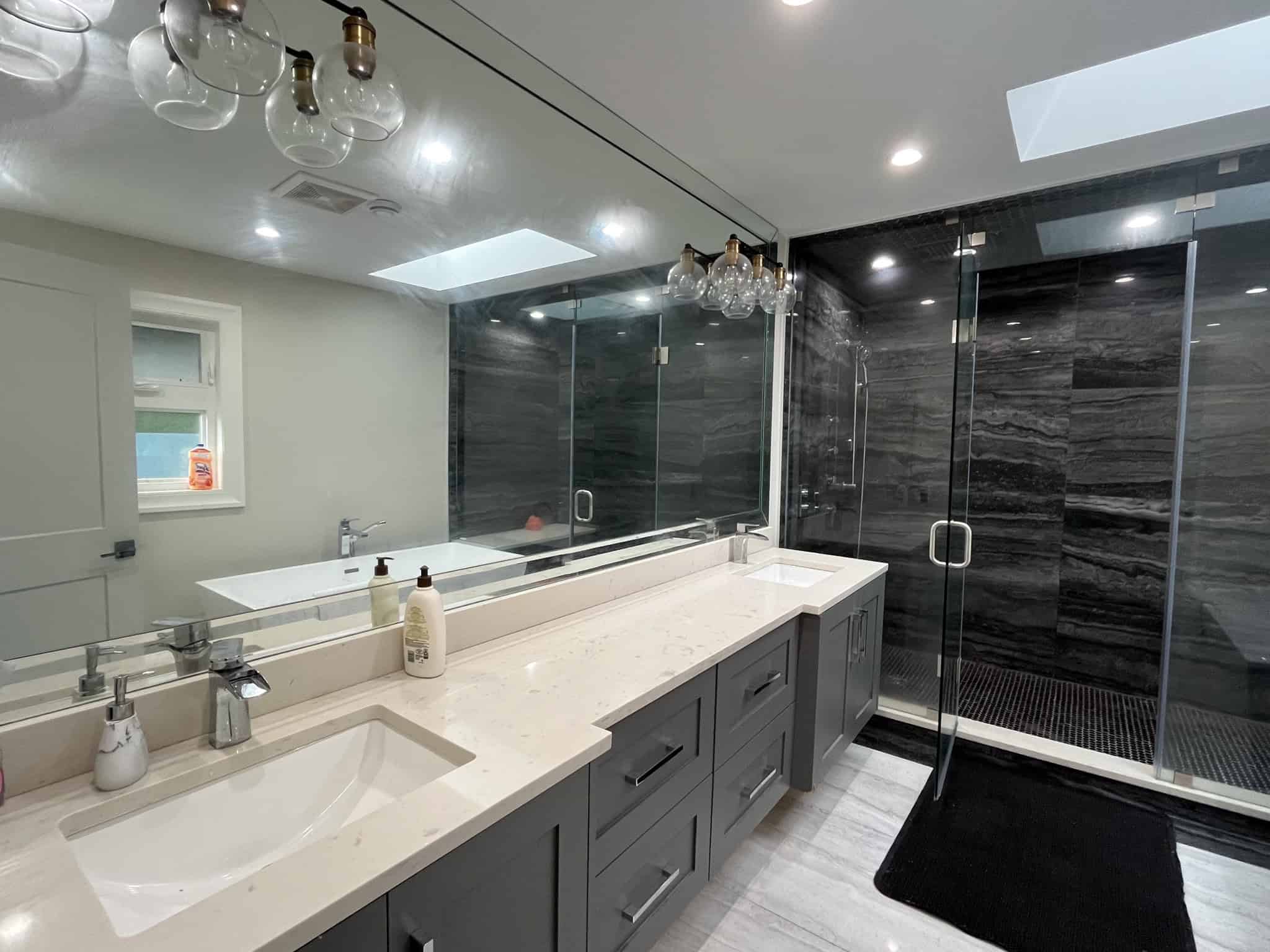 Modern bathroom with a double vanity, marble countertop, and sleek grey cabinetry, featuring a spacious walk-in shower with black stone tiles and glass doors, illuminated by ceiling lights and a skylight.