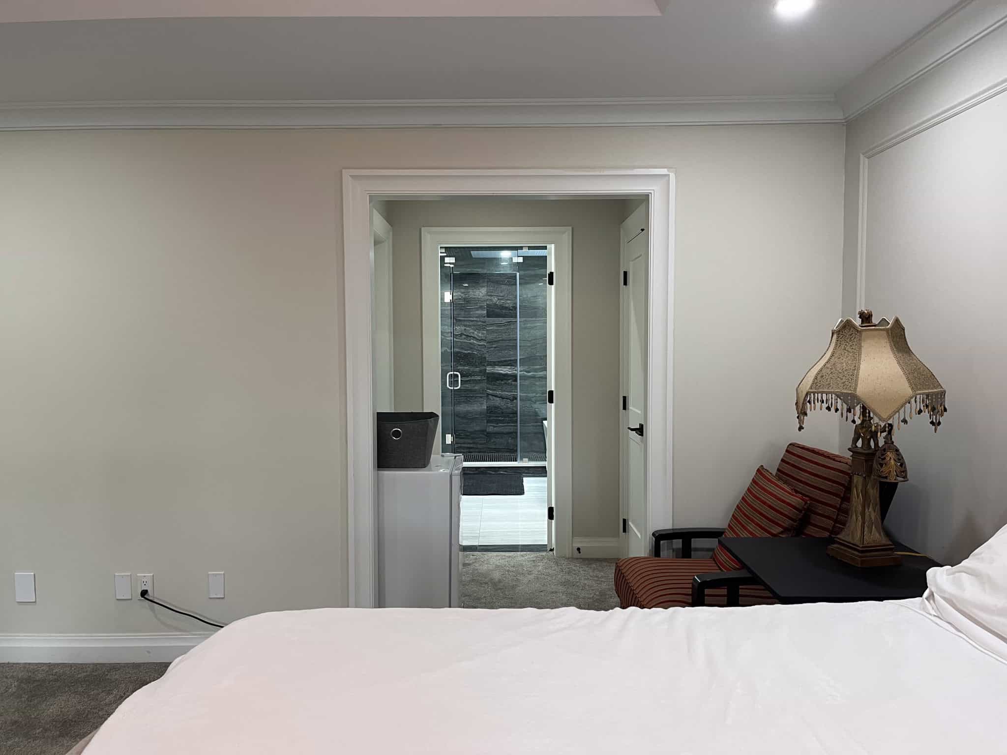 Modern bedroom with a view into a stylish bathroom, featuring elegant decor, a vintage lamp, and a cozy armchair, showcasing upscale interior design and comfort at Orca Realty Inc.