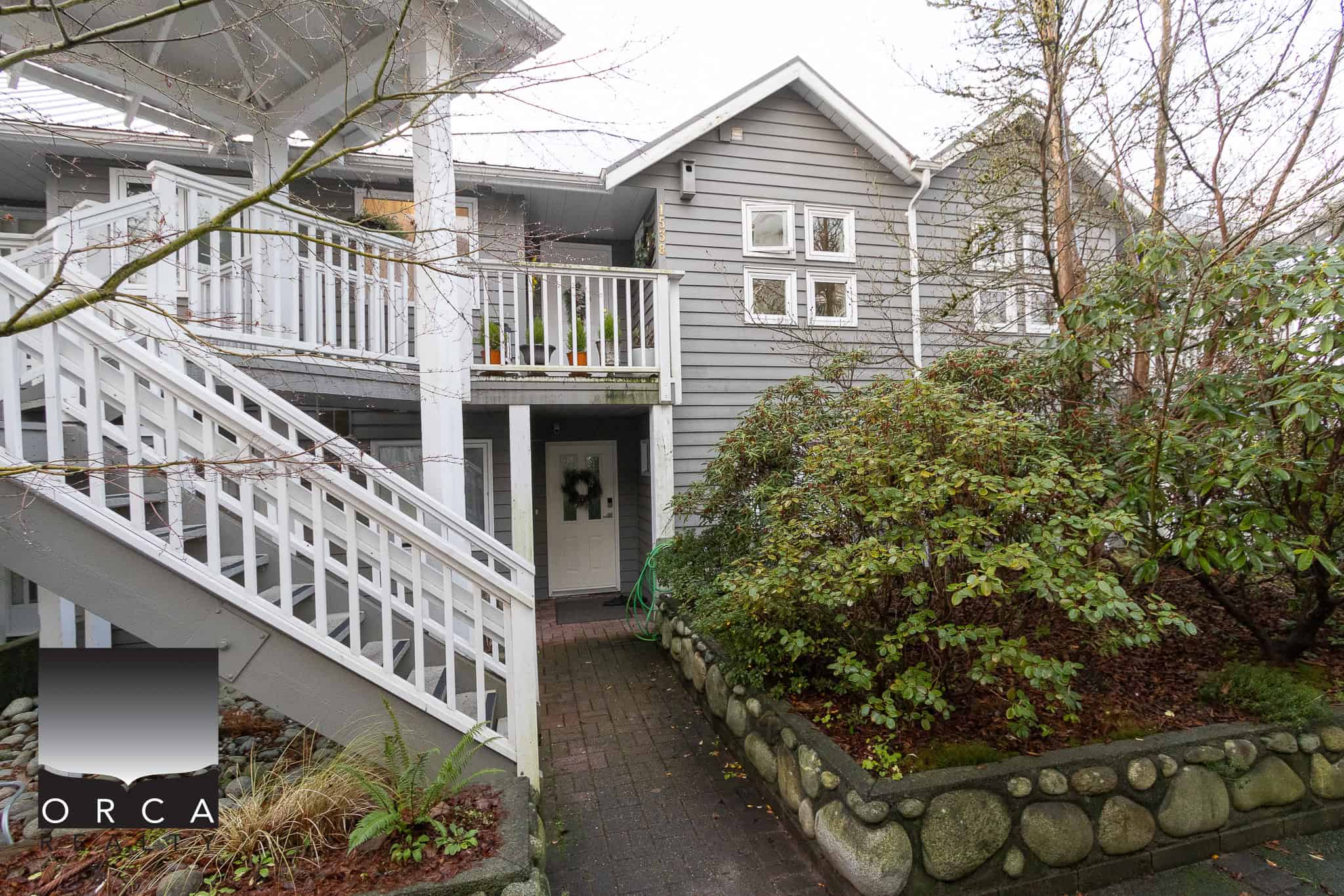 Cozy grey two-story house with white wooden staircase and lush green landscaping, perfect for Vancouver Island homes, available through Orca Realty Inc.