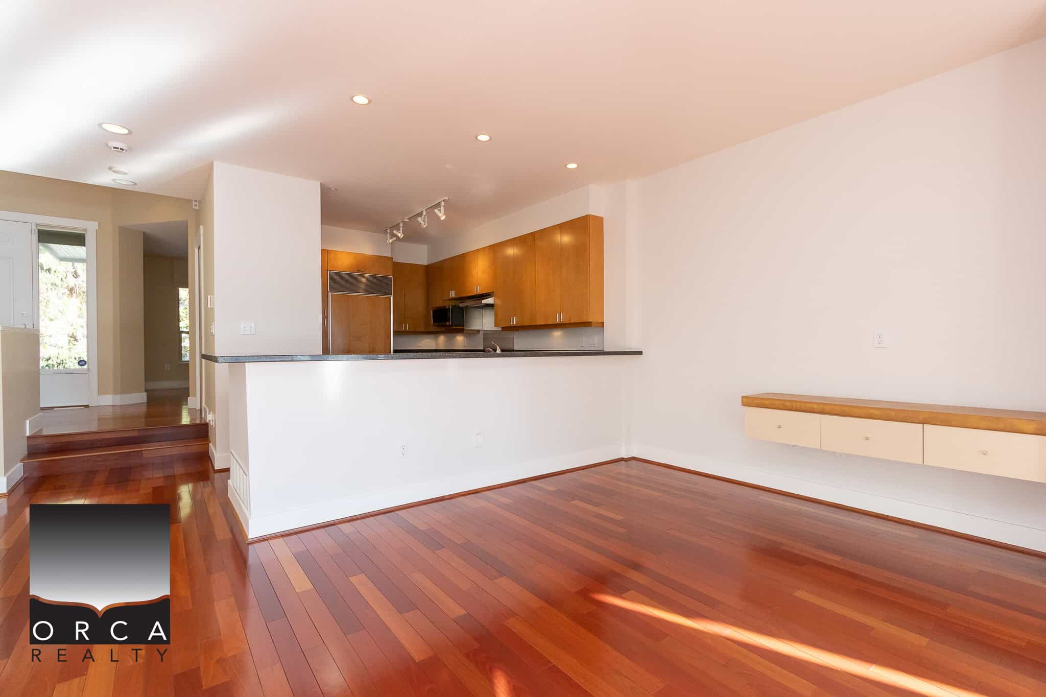 Spacious modern living room with hardwood floors, open kitchen with wooden cabinets, and ample natural light in a contemporary Orca Realty property.