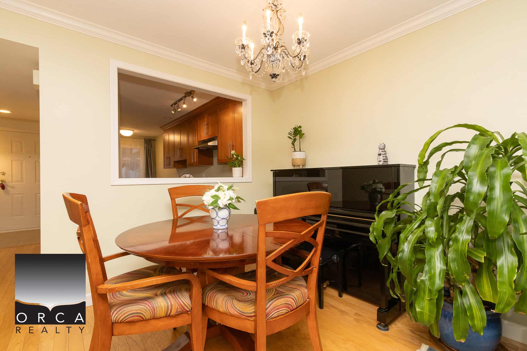 Bright dining room with a wooden table, upholstered chairs, a black upright piano, and vibrant green houseplants, adjacent to a modern kitchen with warm wood cabinetry - perfect for family gatherings.
