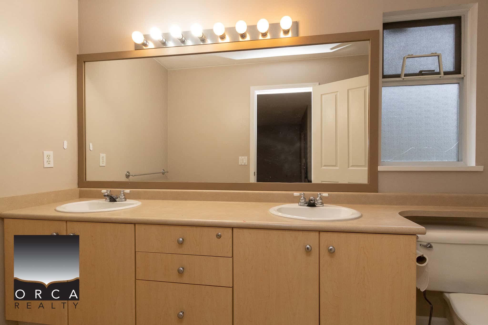 Bright bathroom vanity with dual sinks, beige countertop, and large mirror with overhead lighting, featuring natural light from frosted window, ideal for Vancouver Island real estate.