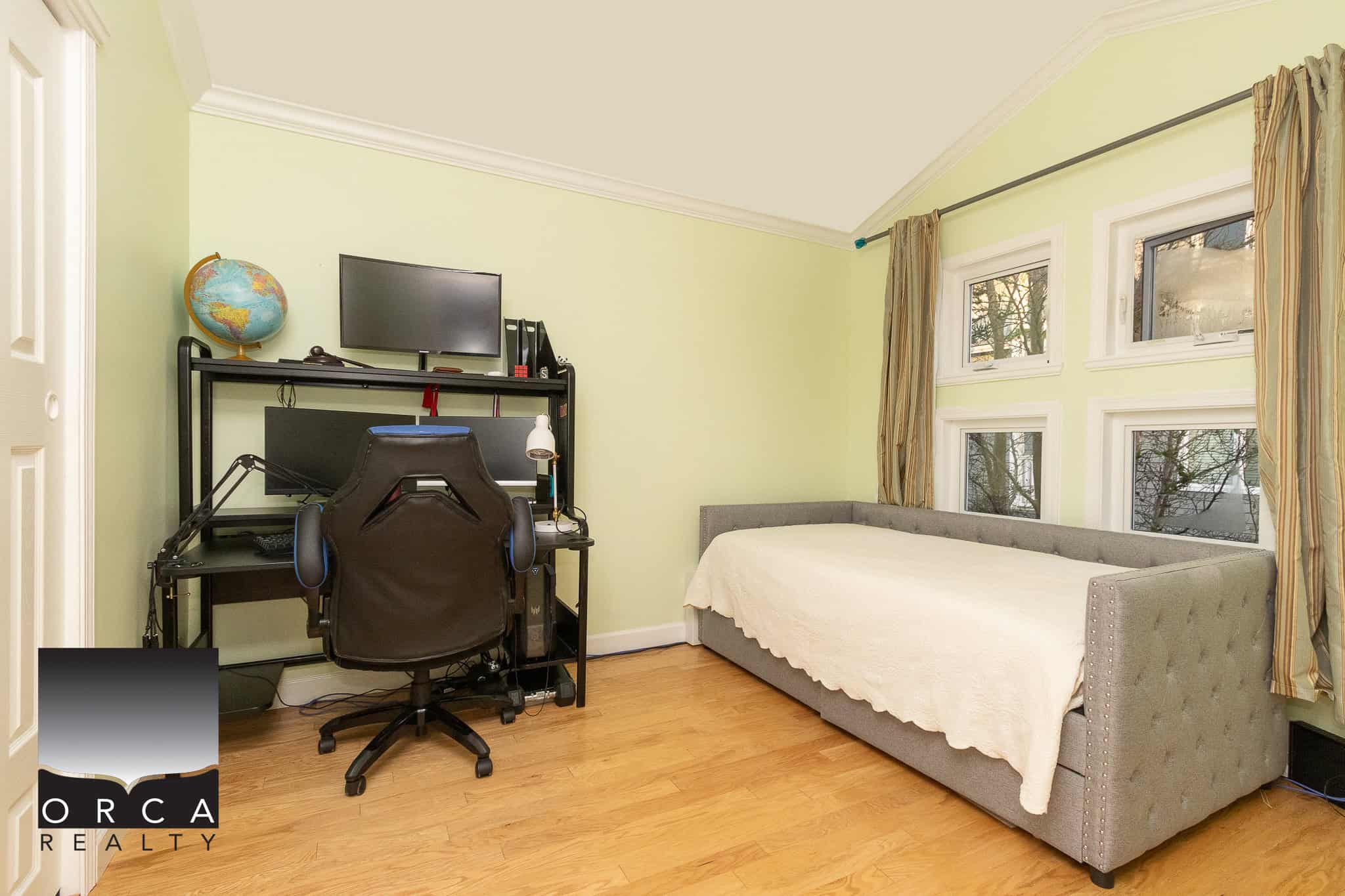 Comfortable home office with a desk, ergonomic chair, computer setup, and a daybed in a bright, sunny room with large windows and light green walls. Ideal for remote work and relaxation.