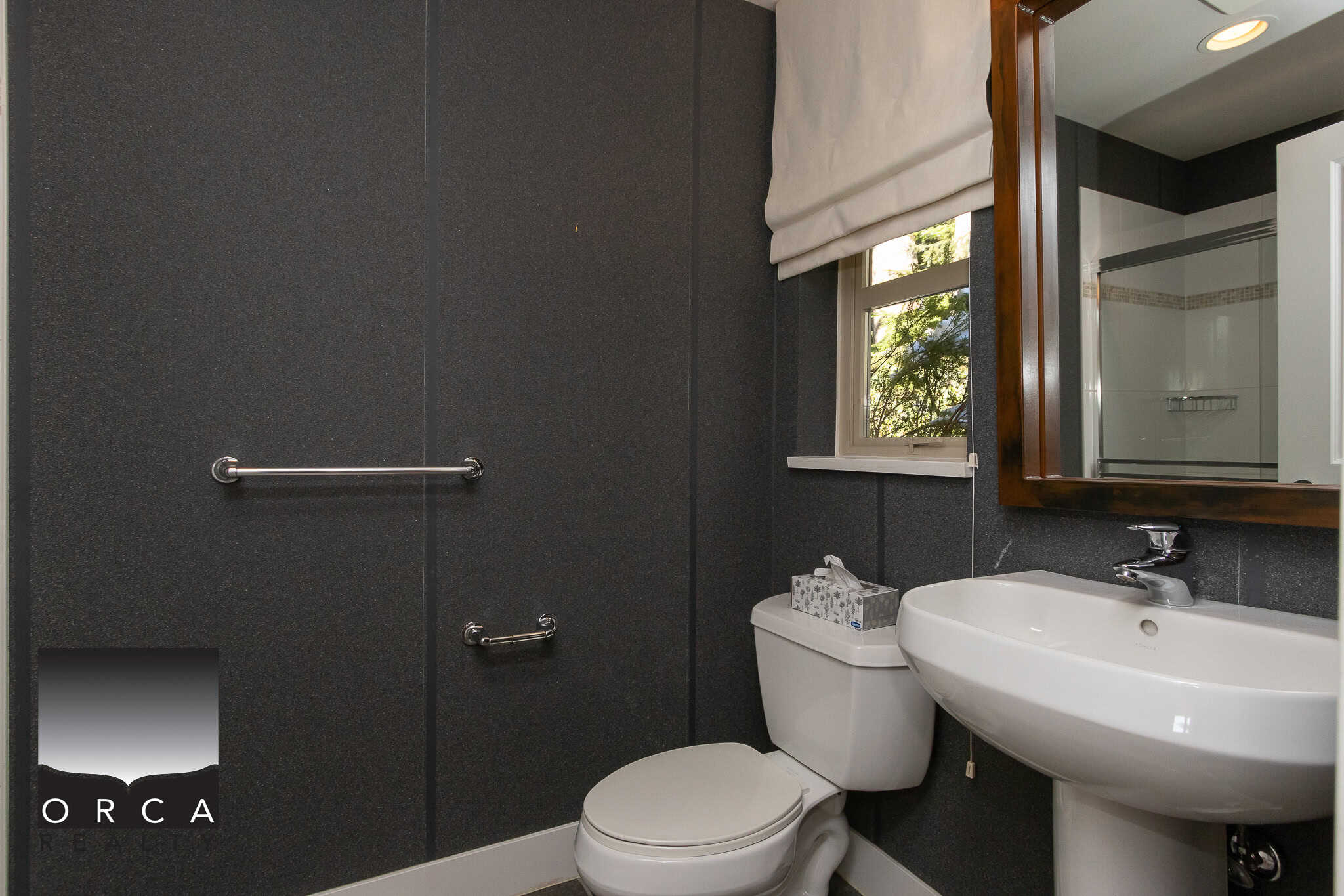 Modern black-textured bathroom with white fixtures, small window with white shade, and dark wood-framed mirror at Orca Realty Inc.