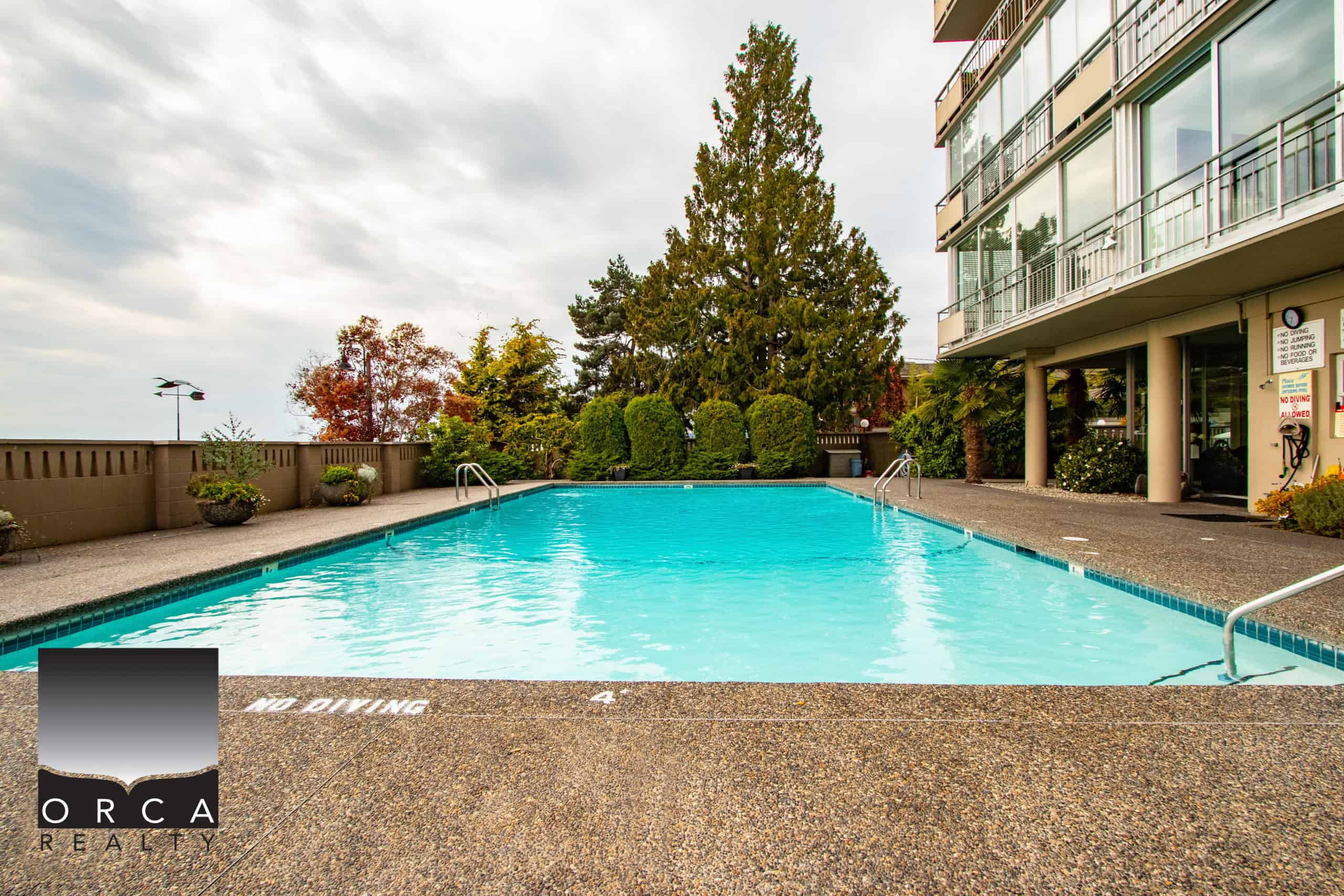 Serene outdoor swimming pool at Orca Realty Inc. property with lush landscaping, adjacent modern residential building, perfect for relaxing and enjoying leisure time in Victoria, BC.