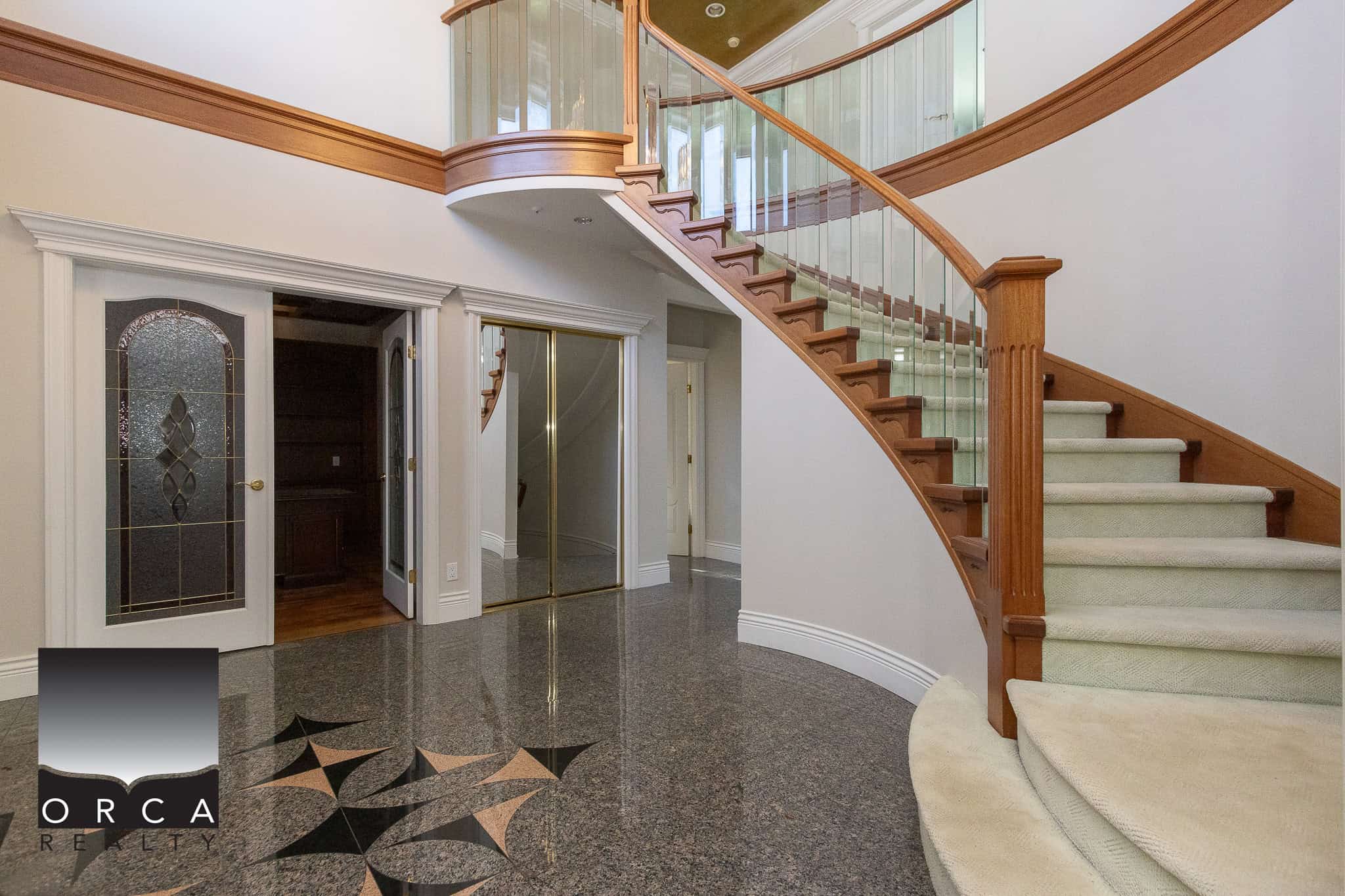 2 Spacious luxury home entrance with elegant curved staircase, glass railing, polished stone flooring, and traditional trim details, showcased by Orca Realty Inc. in Victoria, BC.