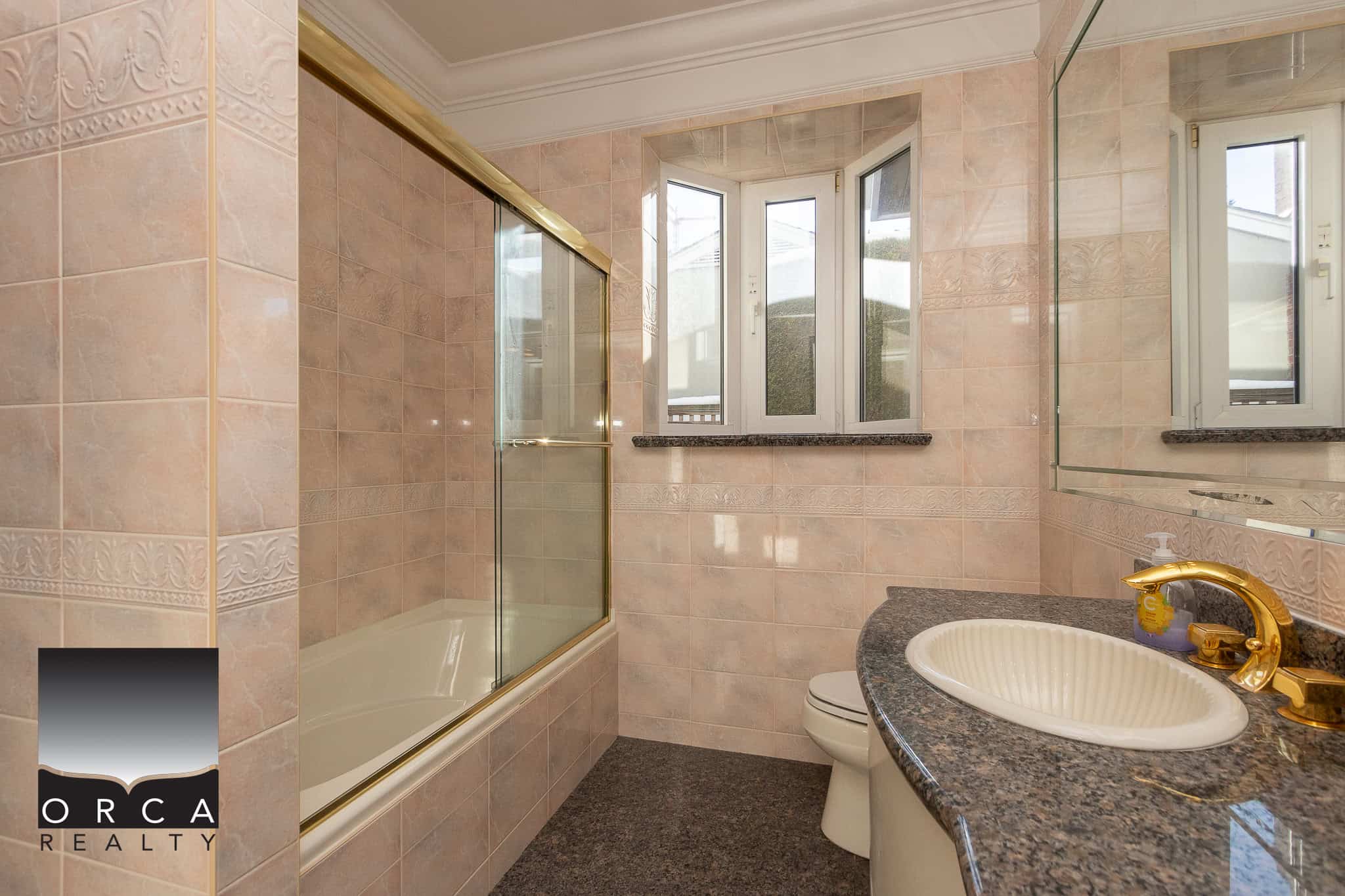20 Bright bathroom with pink ceramic tiles, bathtub with glass shower door, double window with natural light, granite countertop with built-in sink, gold fixtures, and mirror, ideal for Vancouver Island homebuyers.