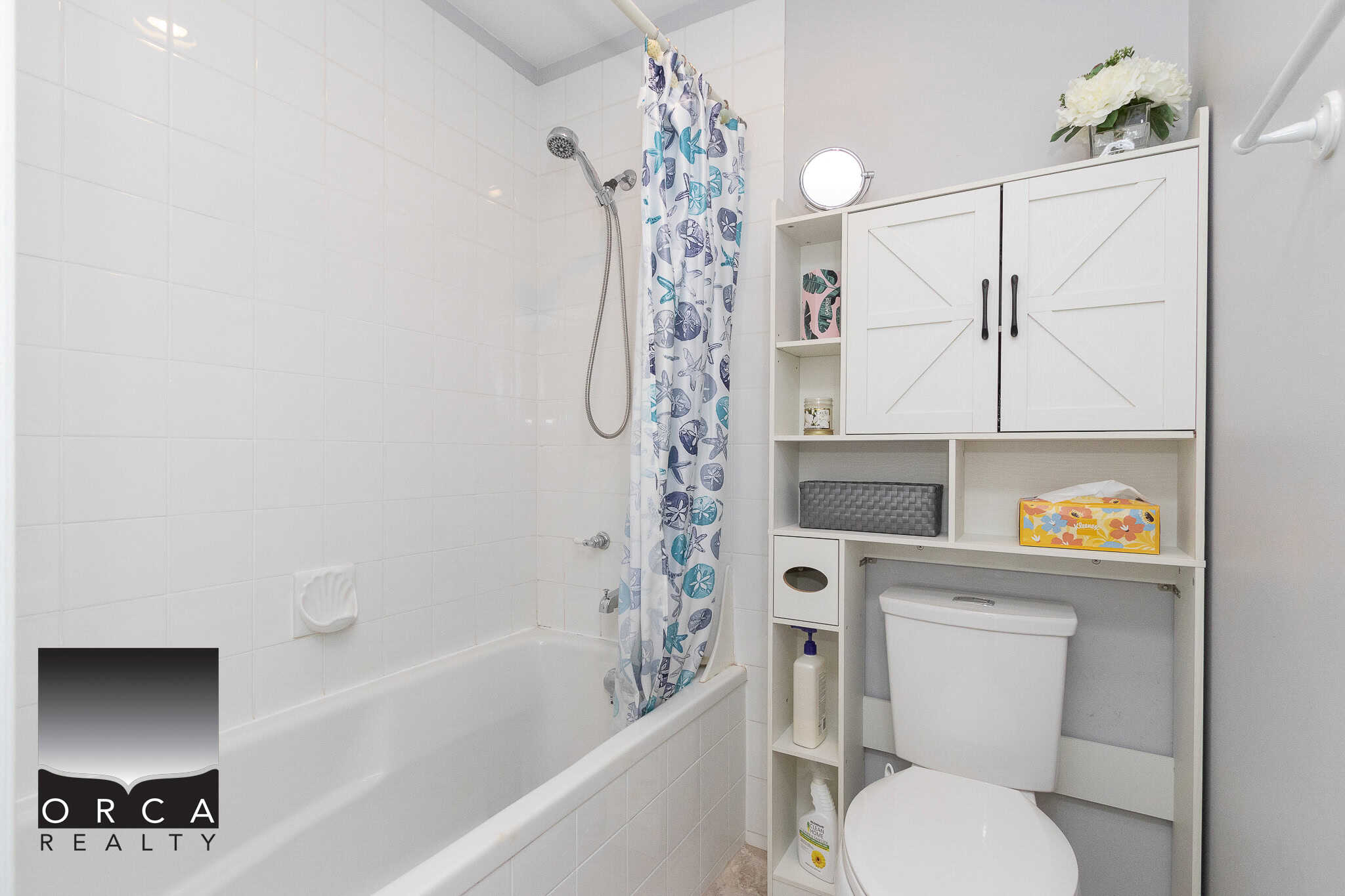 Modern bathroom with white tiled bathtub, decorative shower curtain, and storage cabinet, featuring a toilet, skincare products, and floral decor, ideal for Vancouver residential real estate.