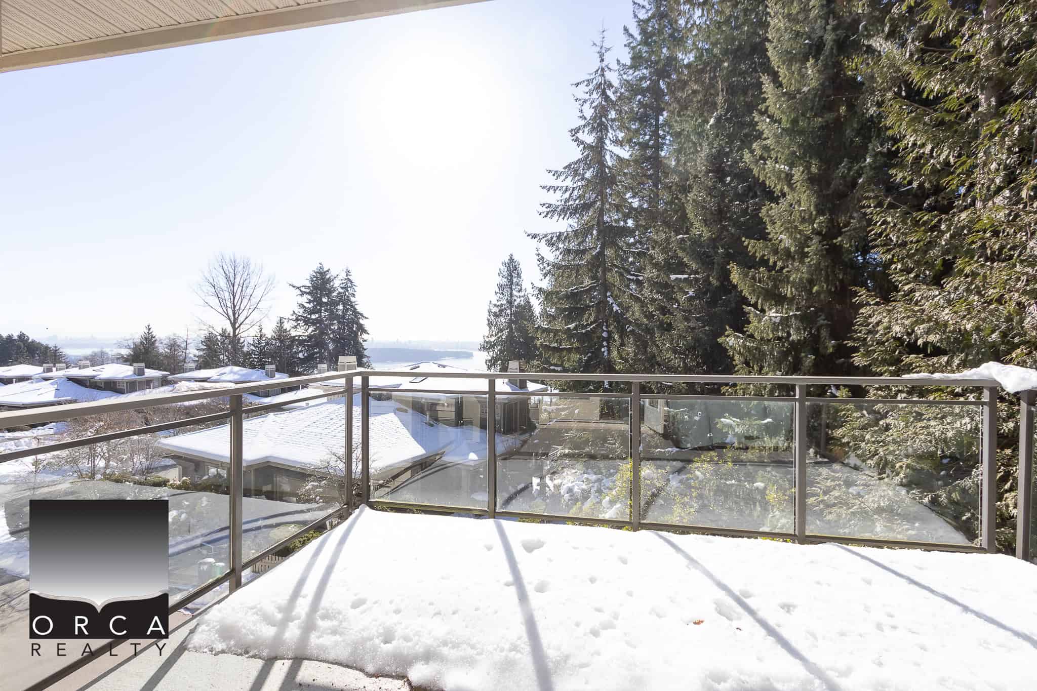 Snow-covered balcony overlooking a serene winter landscape with tall evergreen trees and distant rooftops, ideal for enjoying outdoor views in Vancouver Island real estate.