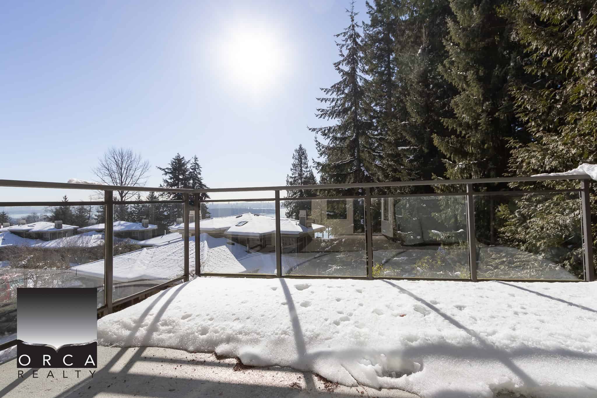 Snow-covered balcony with glass railing overlooking a winter landscape of rooftops and tall evergreen trees, capturing a serene outdoor space in a snowy neighbourhood.