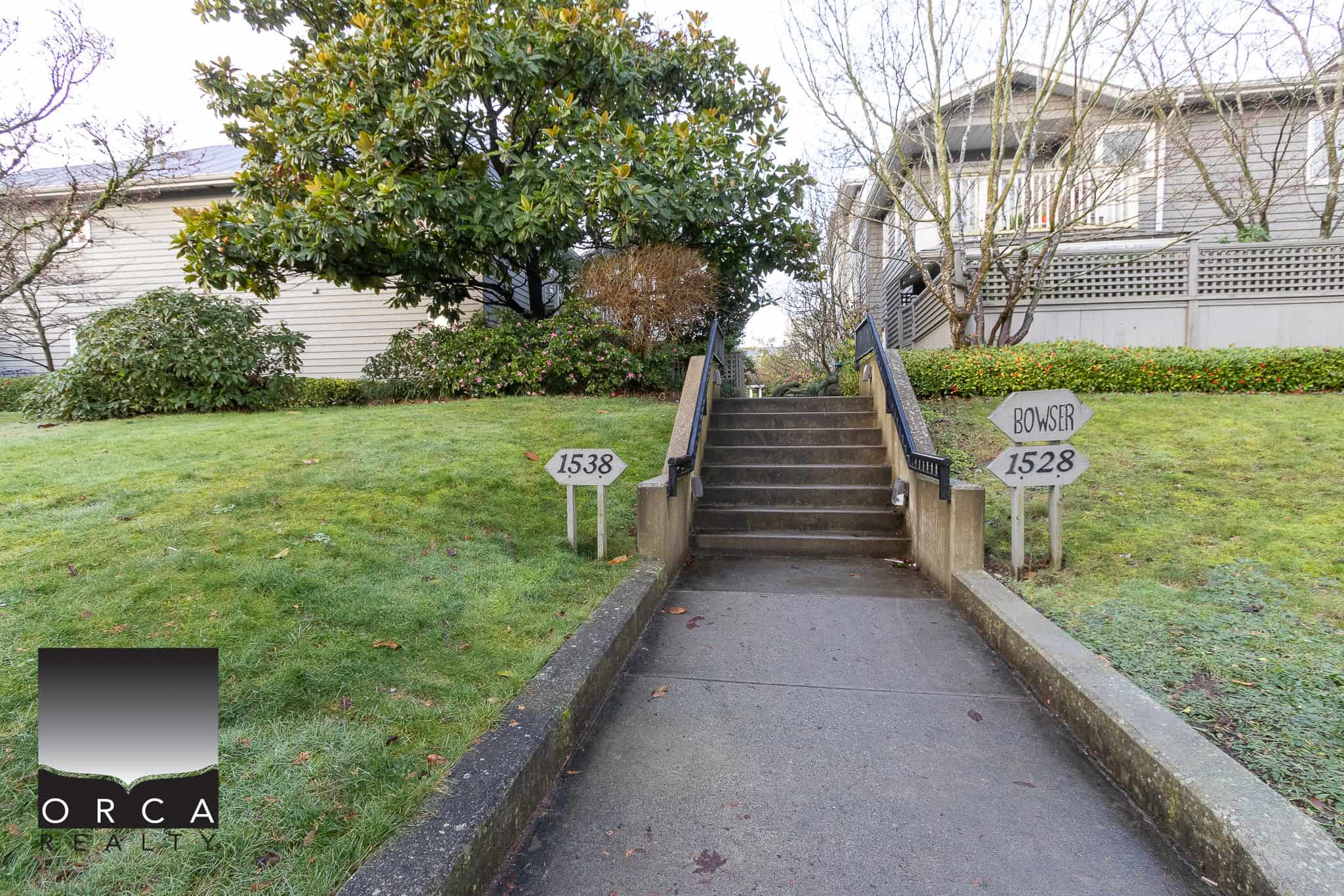 Elevated residential property entrance in a lush, green yard with staircase, surrounded by trees and bushes, showing the inviting nature of homes listed by Orca Realty Inc., experts in Vancouver Island real estate.