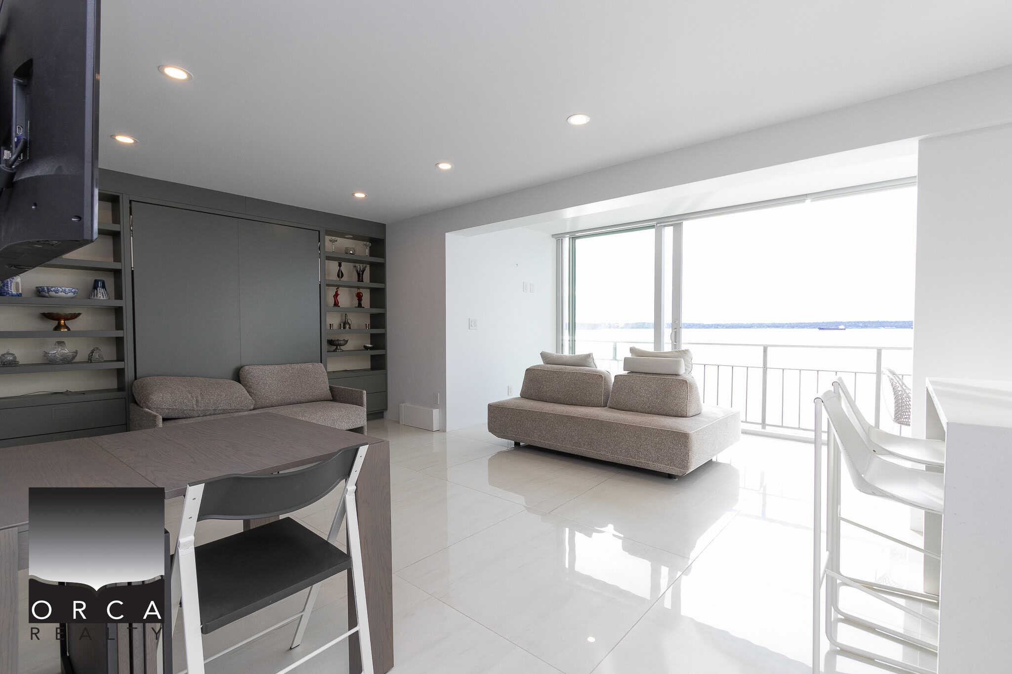 Bright modern living room with ocean views, featuring stylish grey furniture, built-in shelves, and a balcony with panoramic water scenery, ideal for luxury BC real estate.