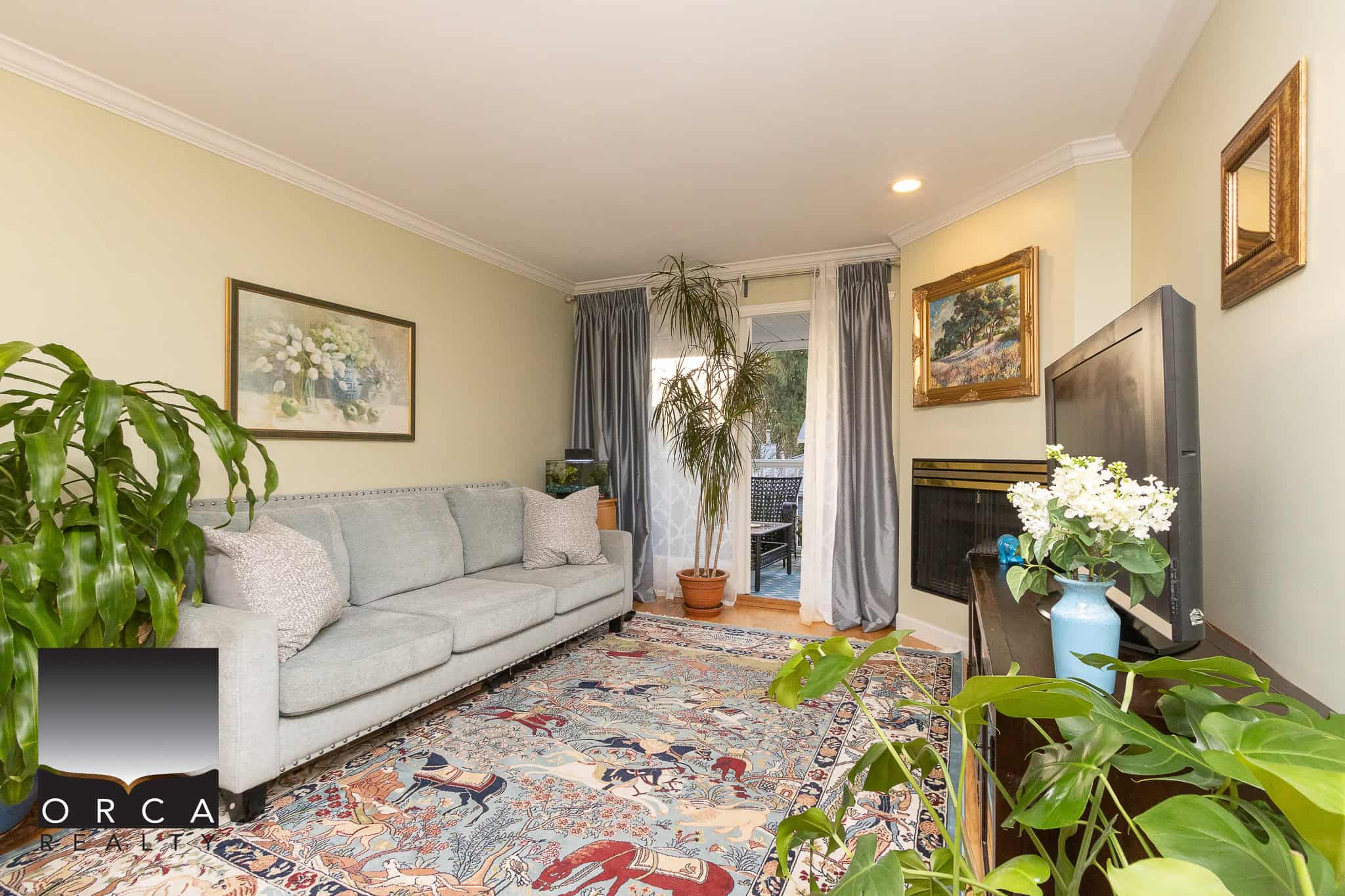 Bright living room with light green walls, comfortable grey sofa, colorful area rug, and potted plants, featuring a balcony door with grey curtains; ideal for Vancouver property shows.