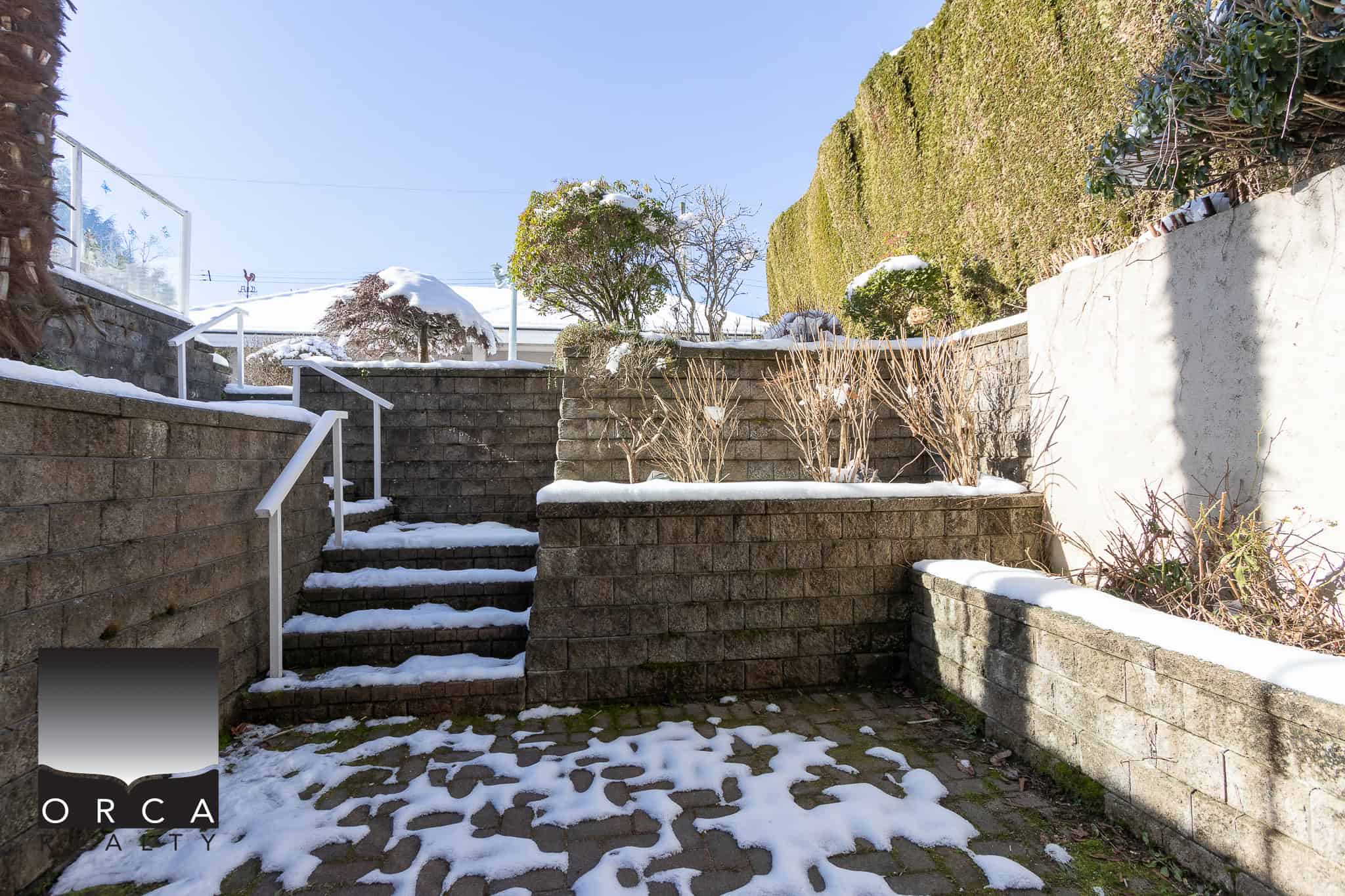 35 Spacious brick backyard patio with snow-covered steps, surrounded by stone walls and lush hedges, ideal for outdoor living in Vancouver.
