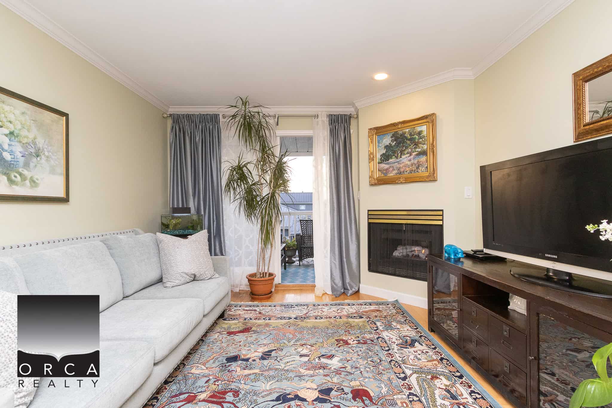 Cozy living room with beige walls, a large grey sofa, colorful area rug, fireplace, and flat-screen TV, featuring elegant artwork and a potted plant, ideal for family gatherings and relaxing evenings, Vancouver real estate.