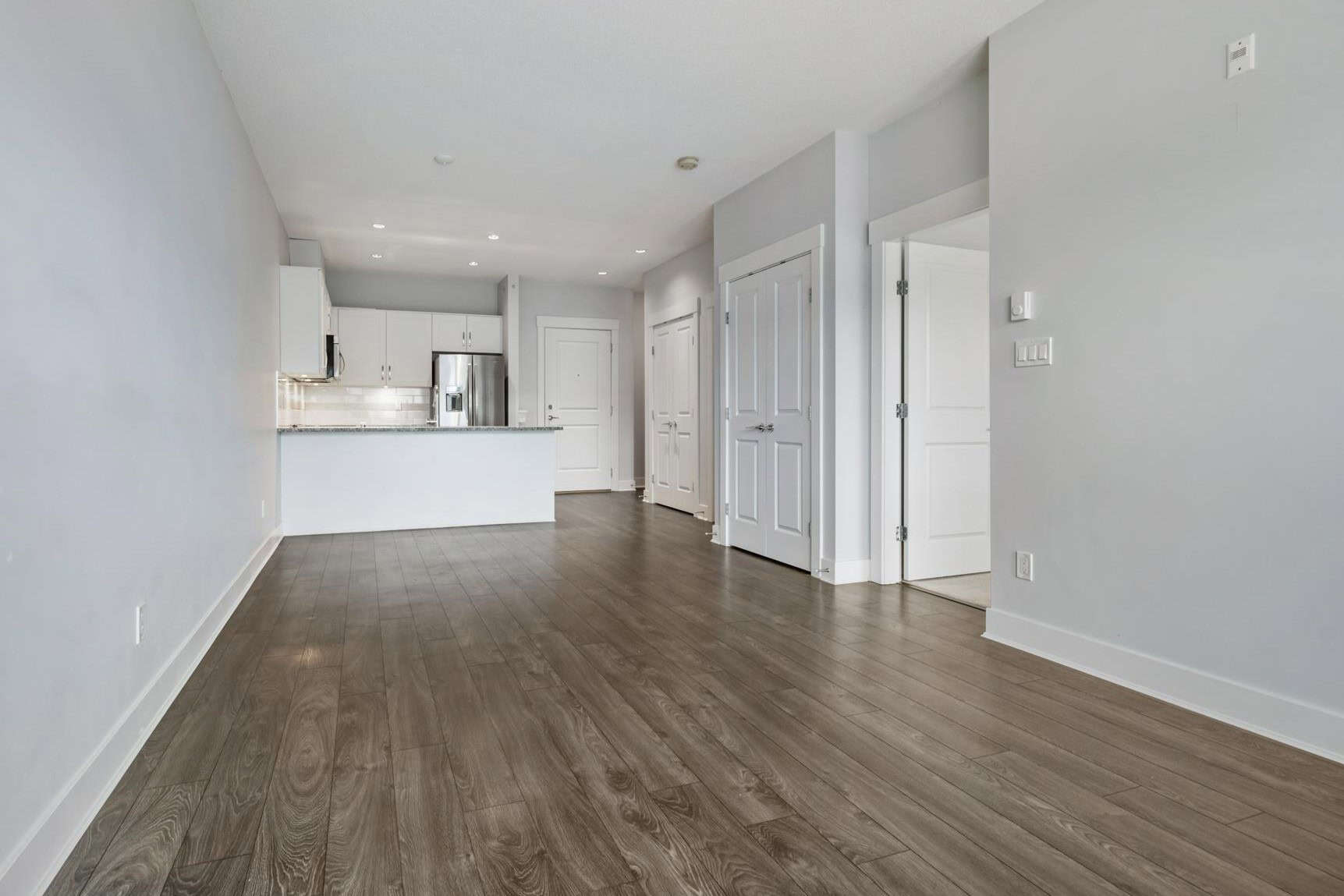 Spacious modern open-concept living room with kitchen, featuring light grey walls, white cabinetry, stainless steel appliances, and wood flooring, ideal for Vancouver real estate.