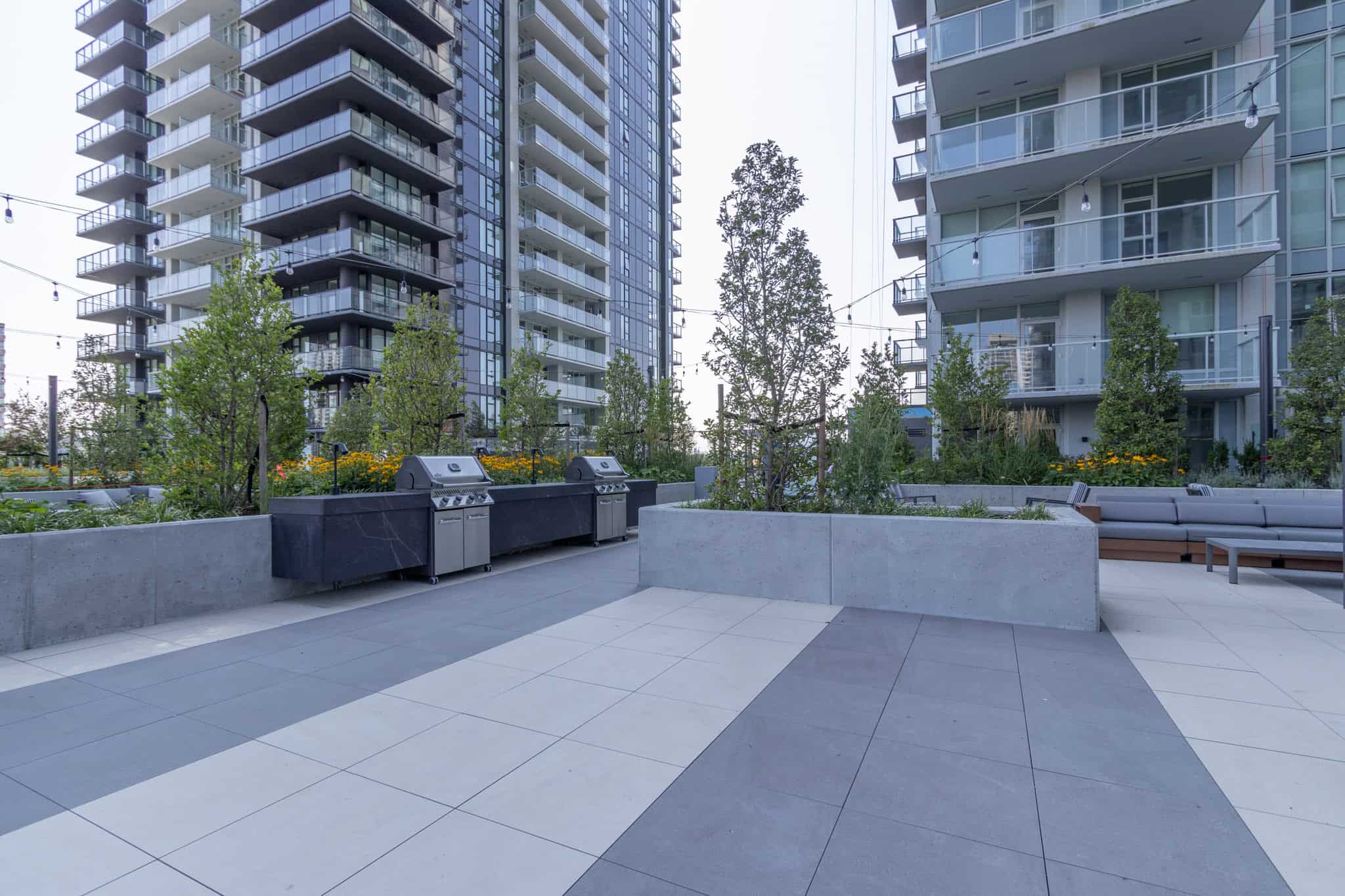 Modern outdoor communal patio area with BBQ grills and seating, surrounded by lush greenery and high-rise residential buildings, showcasing prime Vancouver real estate and amenities.