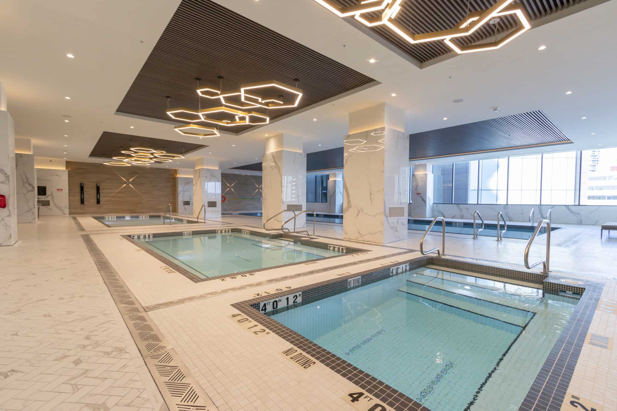 Luxury indoor swimming pool with multiple hot tubs in upscale condo building, modern design, large windows, and elegant lighting.