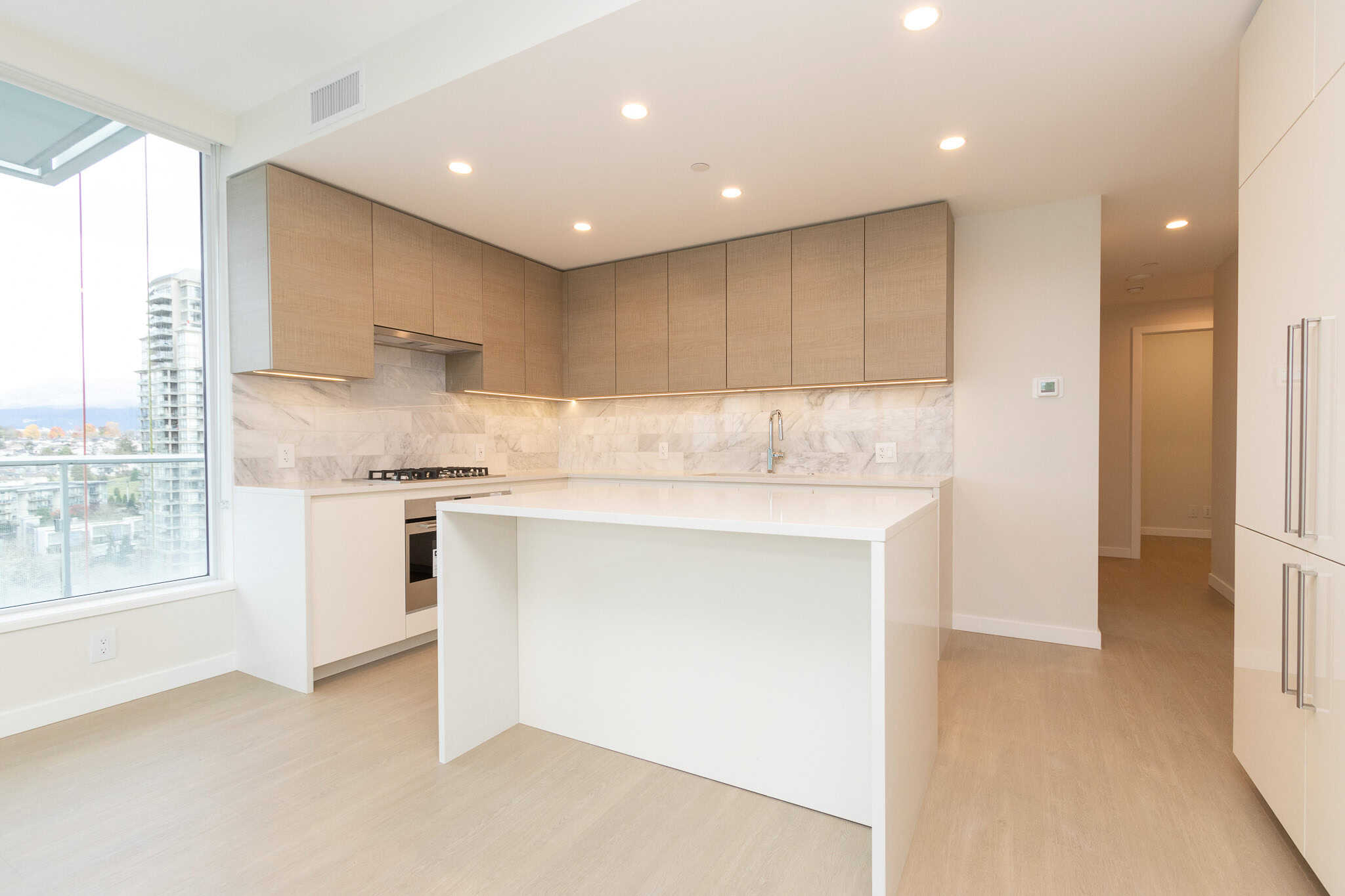 Modern minimalist kitchen with light wood cabinetry, white marble backsplash, and large window showcasing city views. Perfect for contemporary Vancouver-style living.