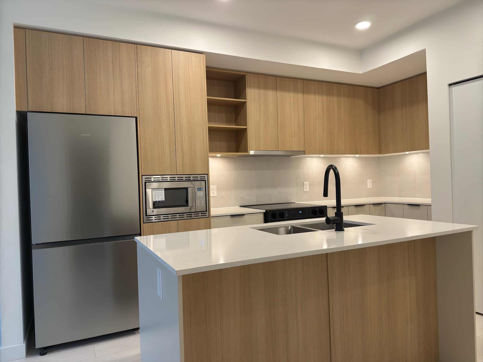 Modern kitchen with light wood cabinetry, stainless steel refrigerator and microwave, black sink faucet, and sleek white countertops, showcasing contemporary design and functionality for potential homebuyers.