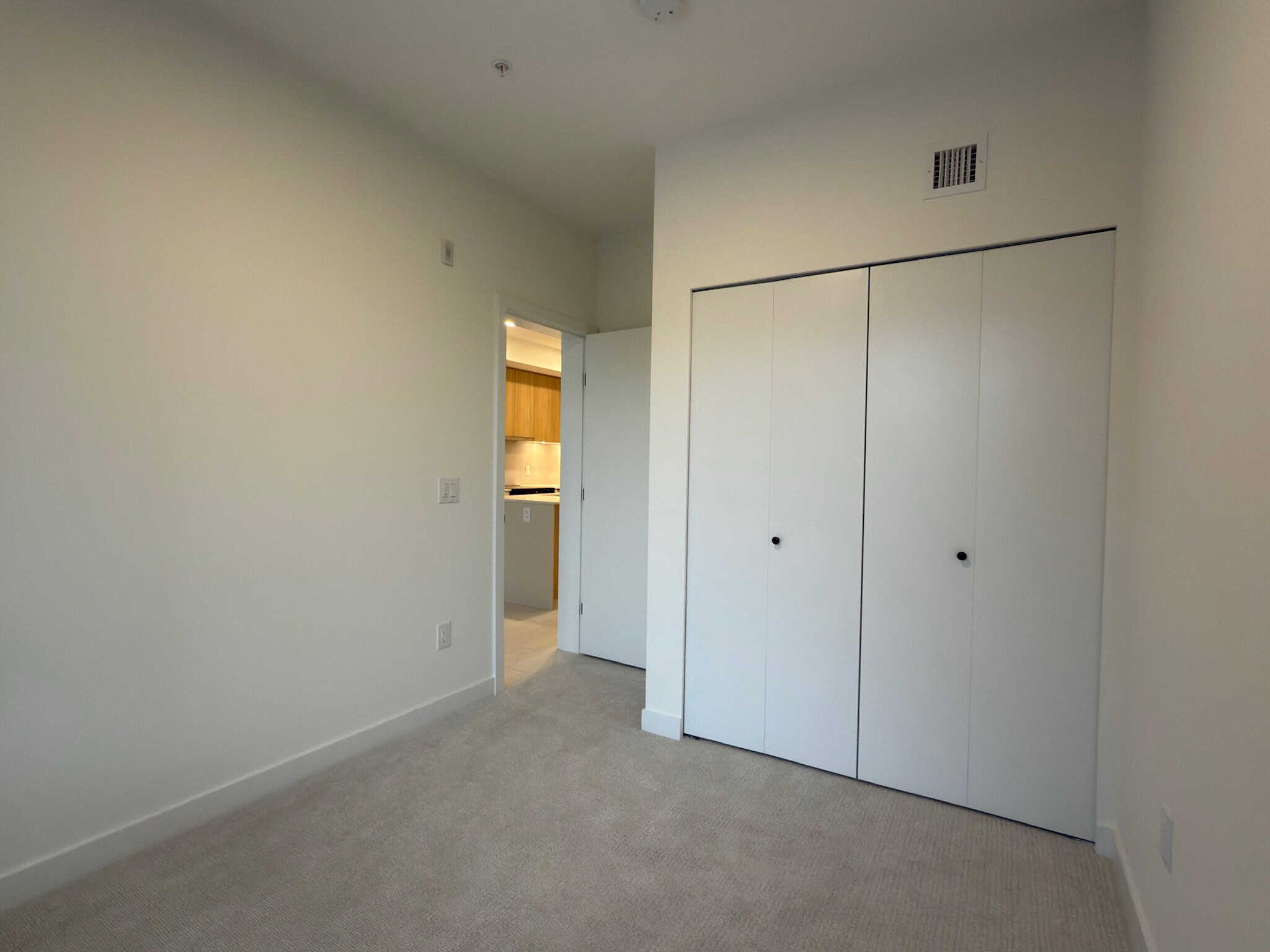 Bright empty bedroom with closet and door to the kitchen, featuring neutral wall paint and carpet flooring, in a property listed by Orca Realty Inc., ideal for residential living in Surrey, BC.