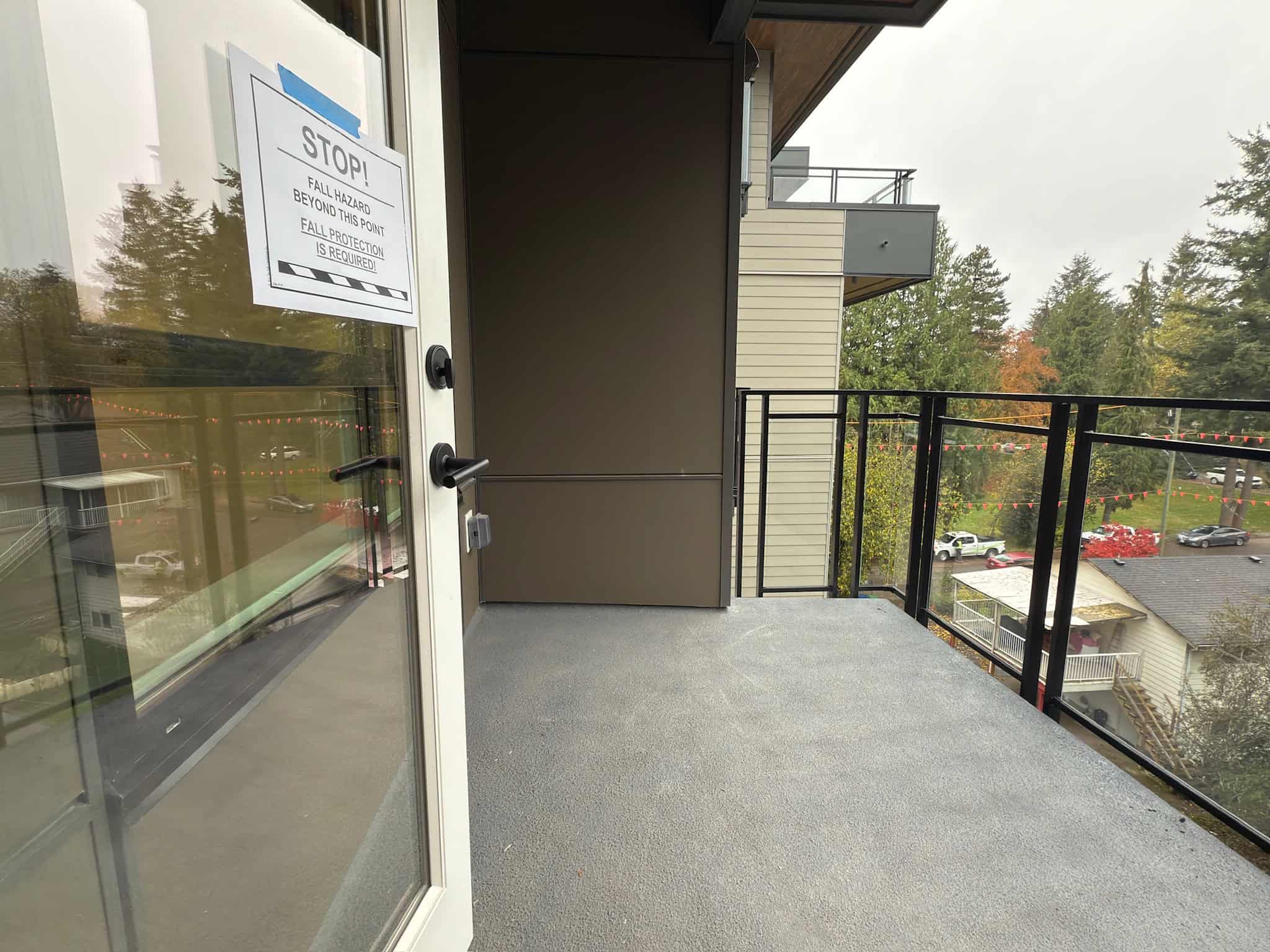 Spacious apartment balcony with safety railing, overlooking greenery and neighboring buildings, featuring an emergency fall hazard warning sign in a residential complex.