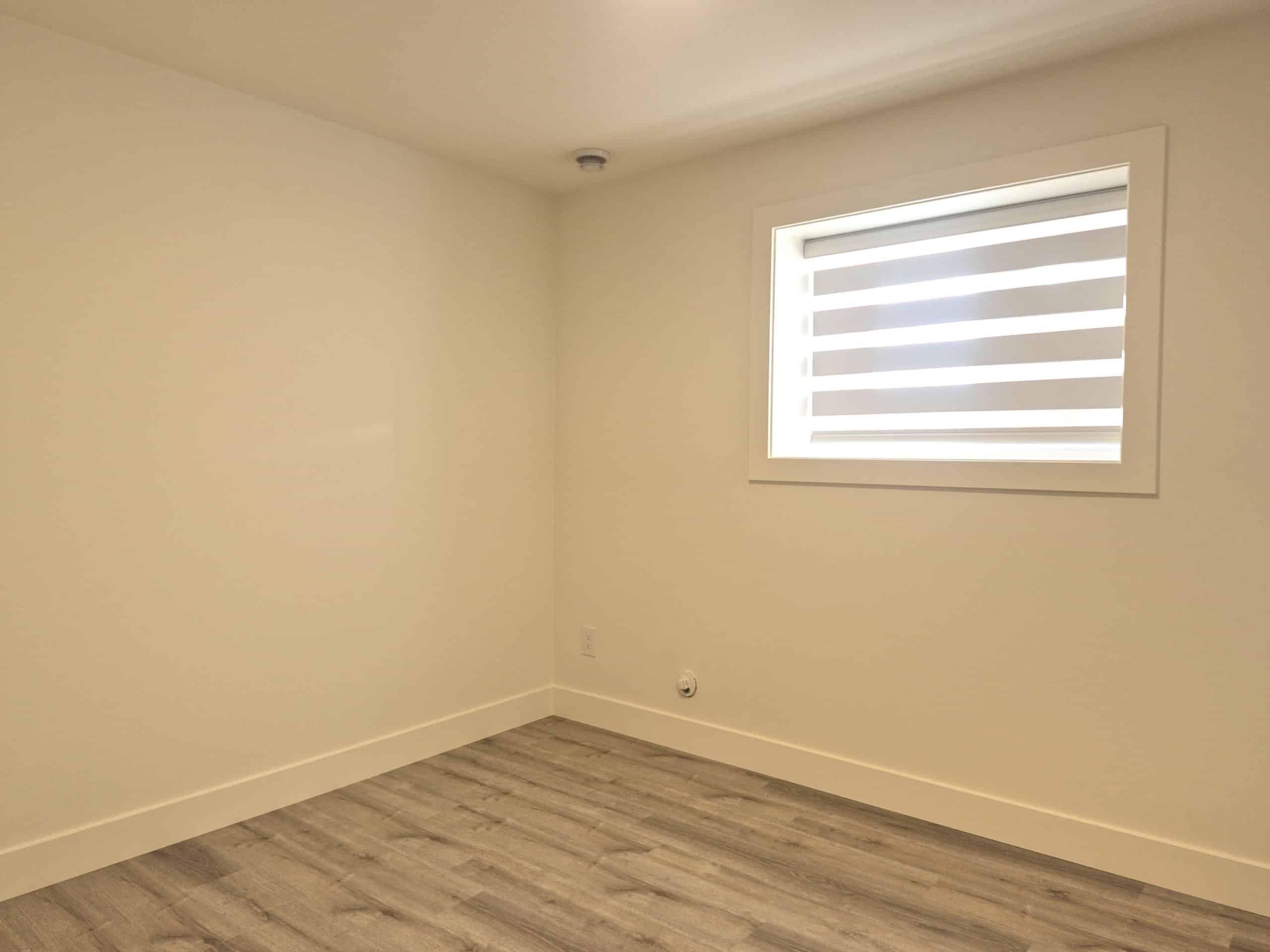 Bright, empty bedroom in a modern home featuring neutral walls, wood-style flooring, and a large window with horizontal blinds, perfect for stable real estate investments or home staging by Orca Realty Inc.