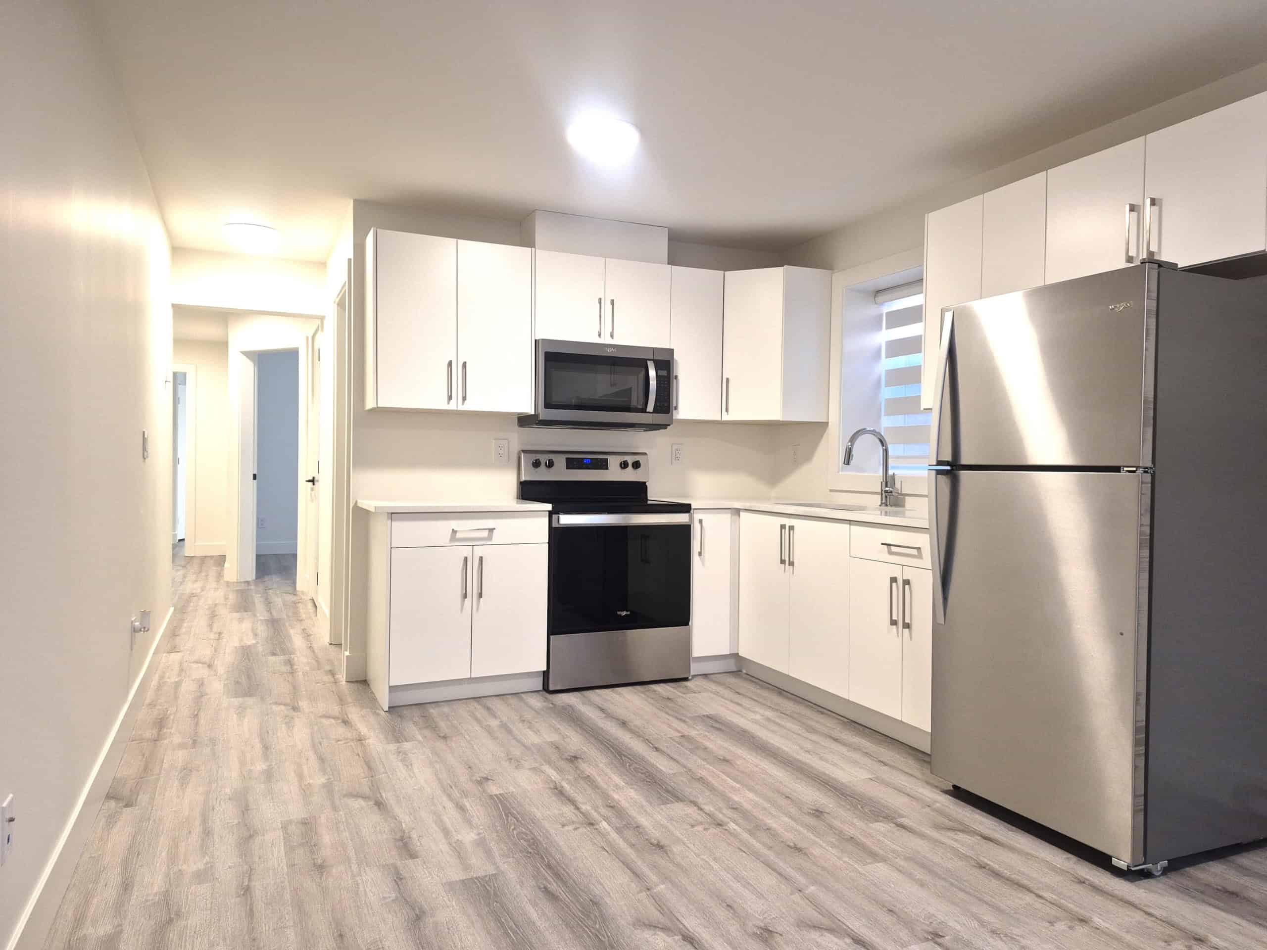 Modern white kitchen with stainless steel appliances, including refrigerator, microwave, and oven, featuring ample cabinetry and light wood flooring for a sleek, contemporary look.