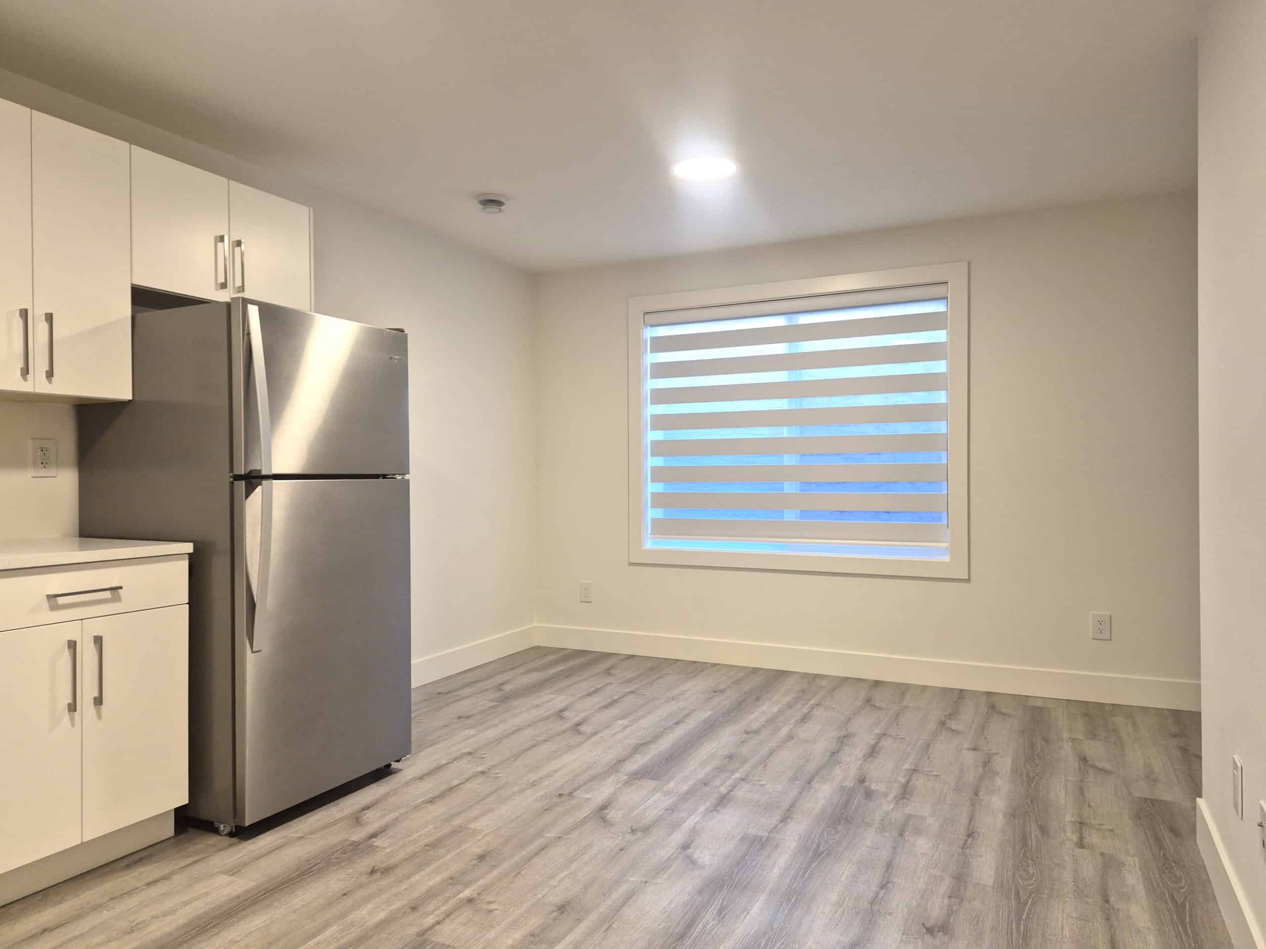 Modern kitchen with white cabinets and stainless steel refrigerator, featuring a large window with horizontal blinds, hardwood flooring, and minimalist design, ideal for Vancouver Real Estate listings.