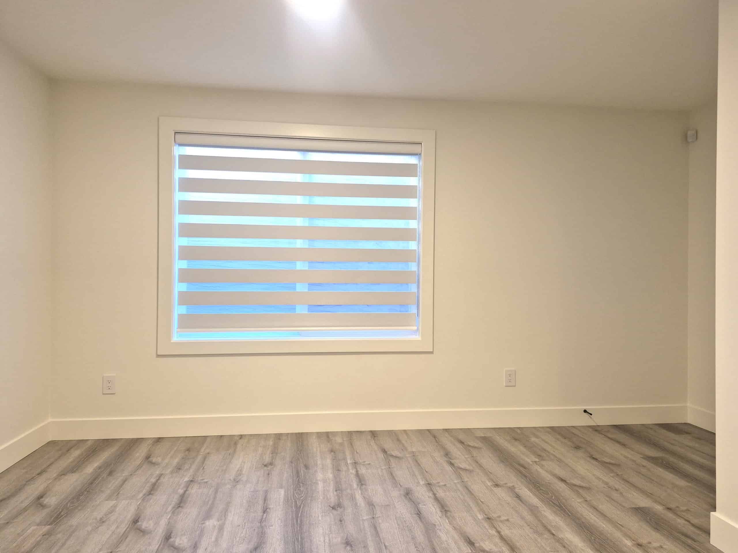 Bright empty room with modern laminate flooring and a large window with zebra blinds, perfect for real estate listings or interior design showcases.