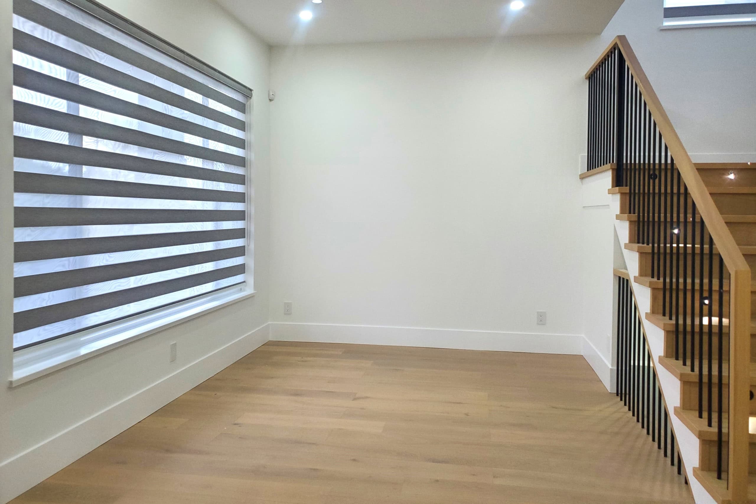 7 Modern interior living space featuring wood flooring, large window with zebra blinds, and a staircase with black metal balusters and wooden handrail, ideal for Vancouver real estate listings.