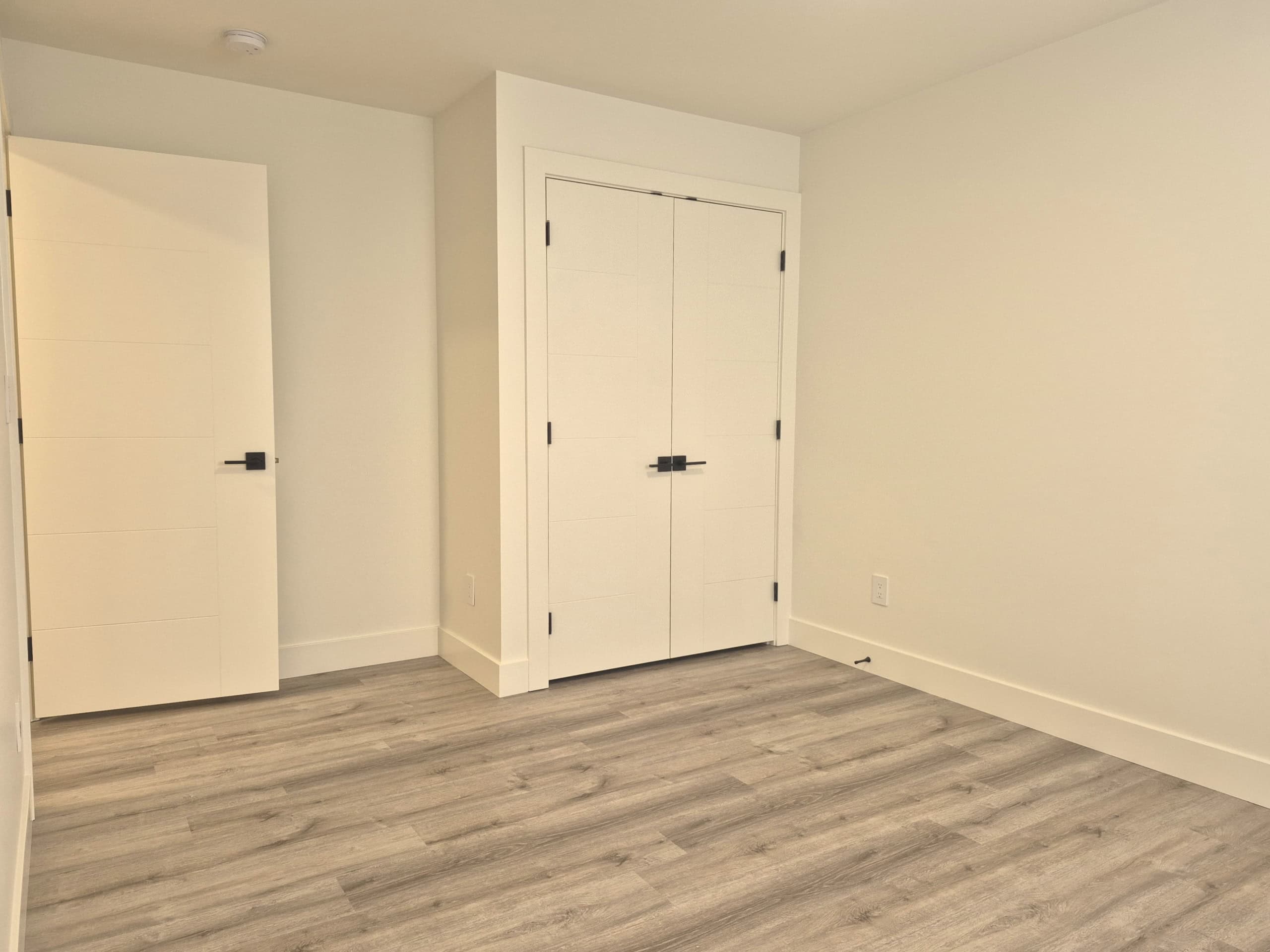Bright, modern bedroom with white walls, sleek black hardware on closet doors, and light grey wood flooring, ideal for a stylish Vancouver home or condo.