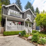 Charming grey Vancouver home with white accents, lush landscaping, and a welcoming front porch, showcasing beautiful garden beds and mature trees in a peaceful neighbourhood.
